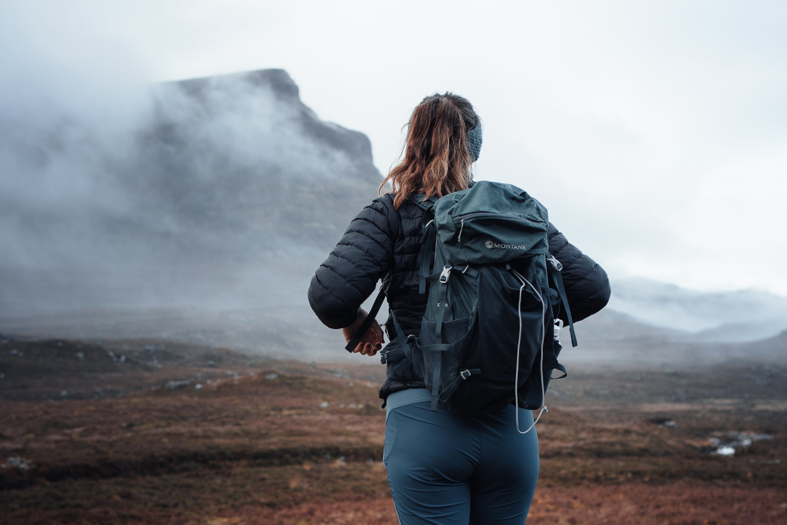 A woman with brown hair wearing a gray mask, dark jacket, and blue hiking pants, carrying a gray backpack, hiking in a foggy, rocky landscape.