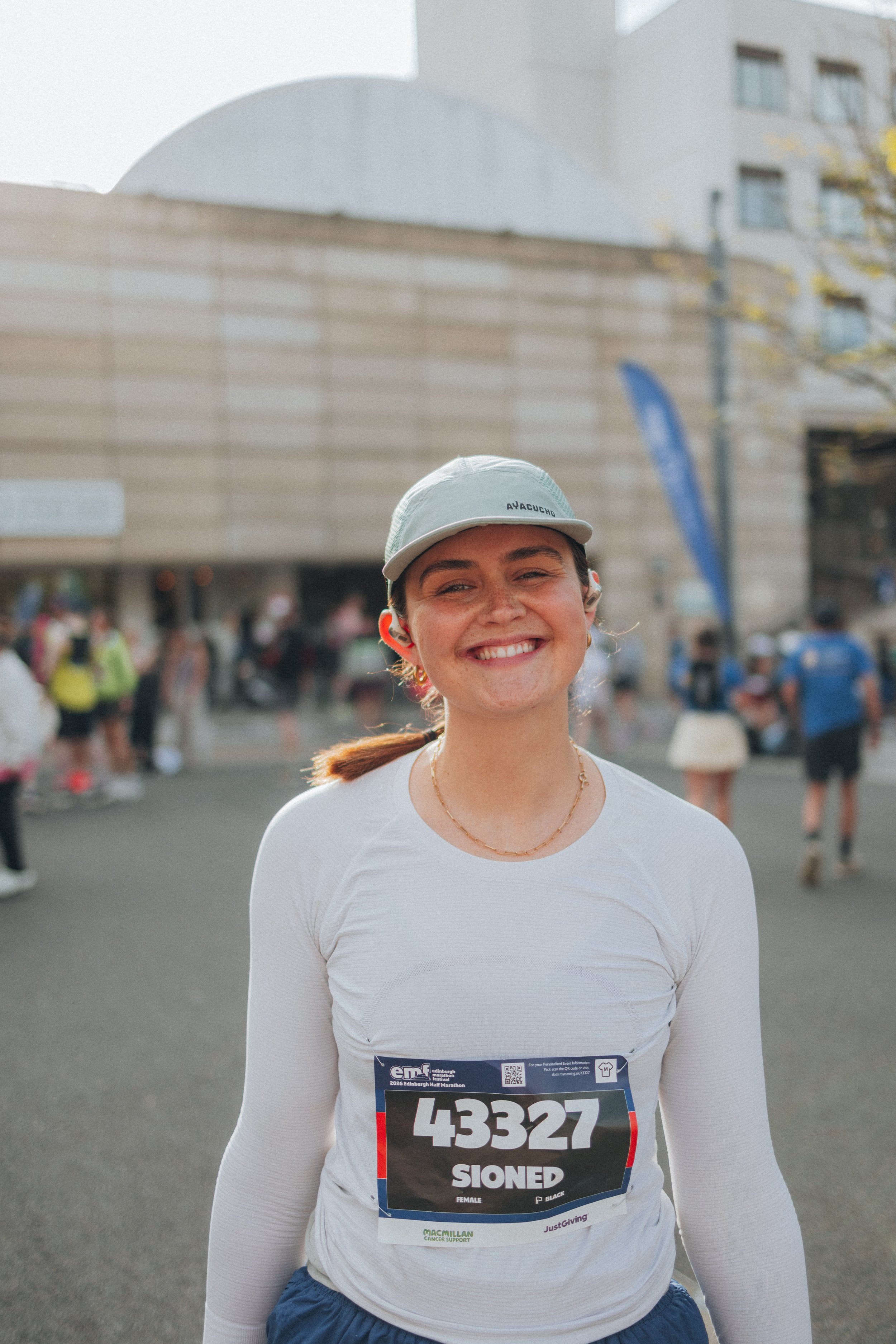A smiling woman wearing a gray cap, white long-sleeve shirt, and a race bib number 43327 at a marathon event outdoors on a sunny day.