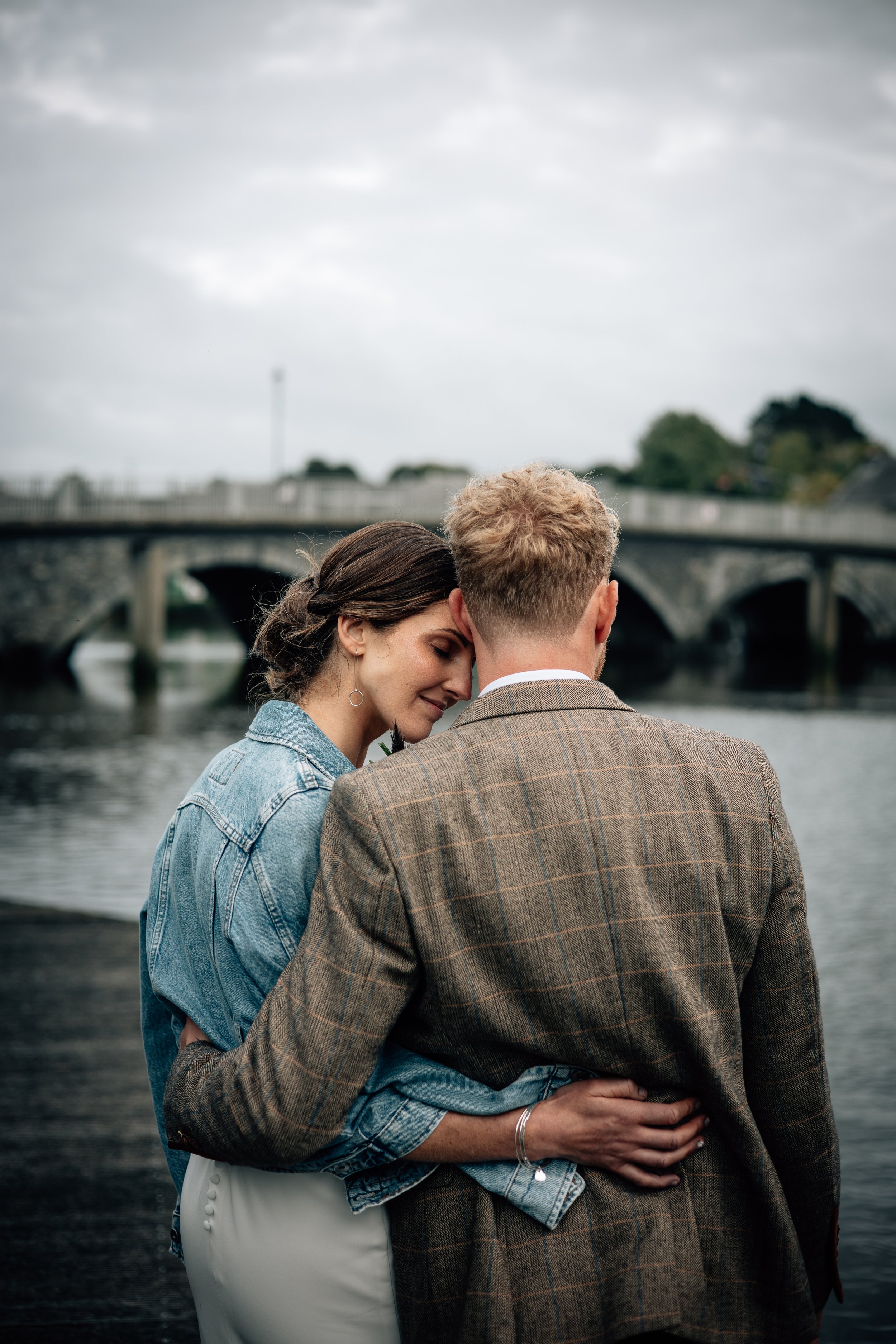 A couple hugging near a river with a bridge in the background on a cloudy day