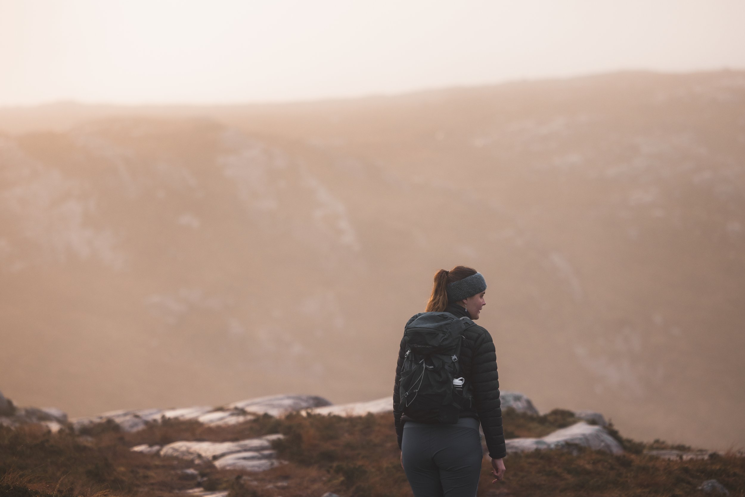 A woman with reddish hair in a ponytail, wearing a black puffy jacket and backpack, stands outdoors on rocky terrain during sunset or sunrise, with a soft, warm sky in the background.