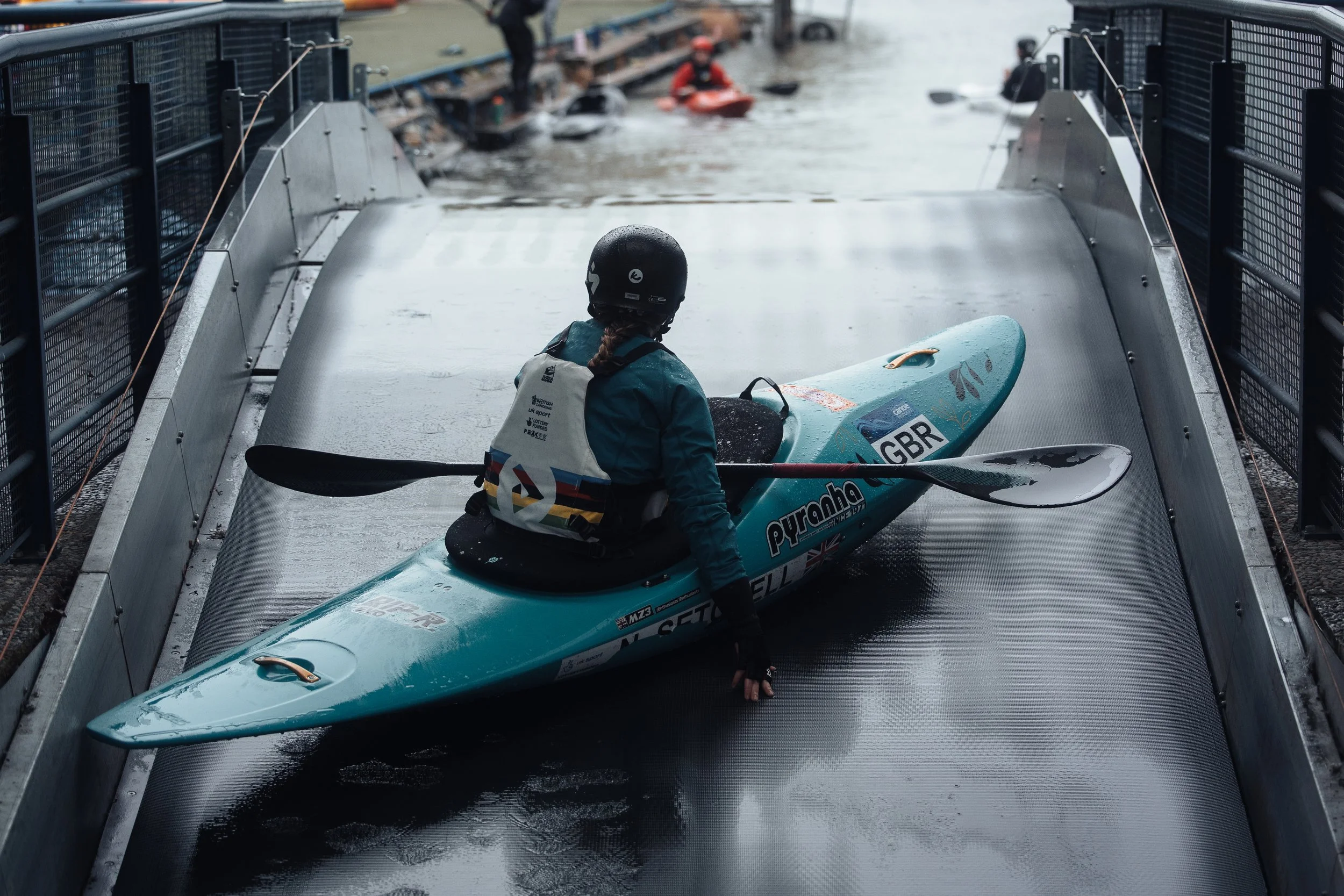 Person in a wetsuit with a helmet and life jacket, holding a kayak, sitting at the bottom of a ramp with water, with other kayakers visible in the background.