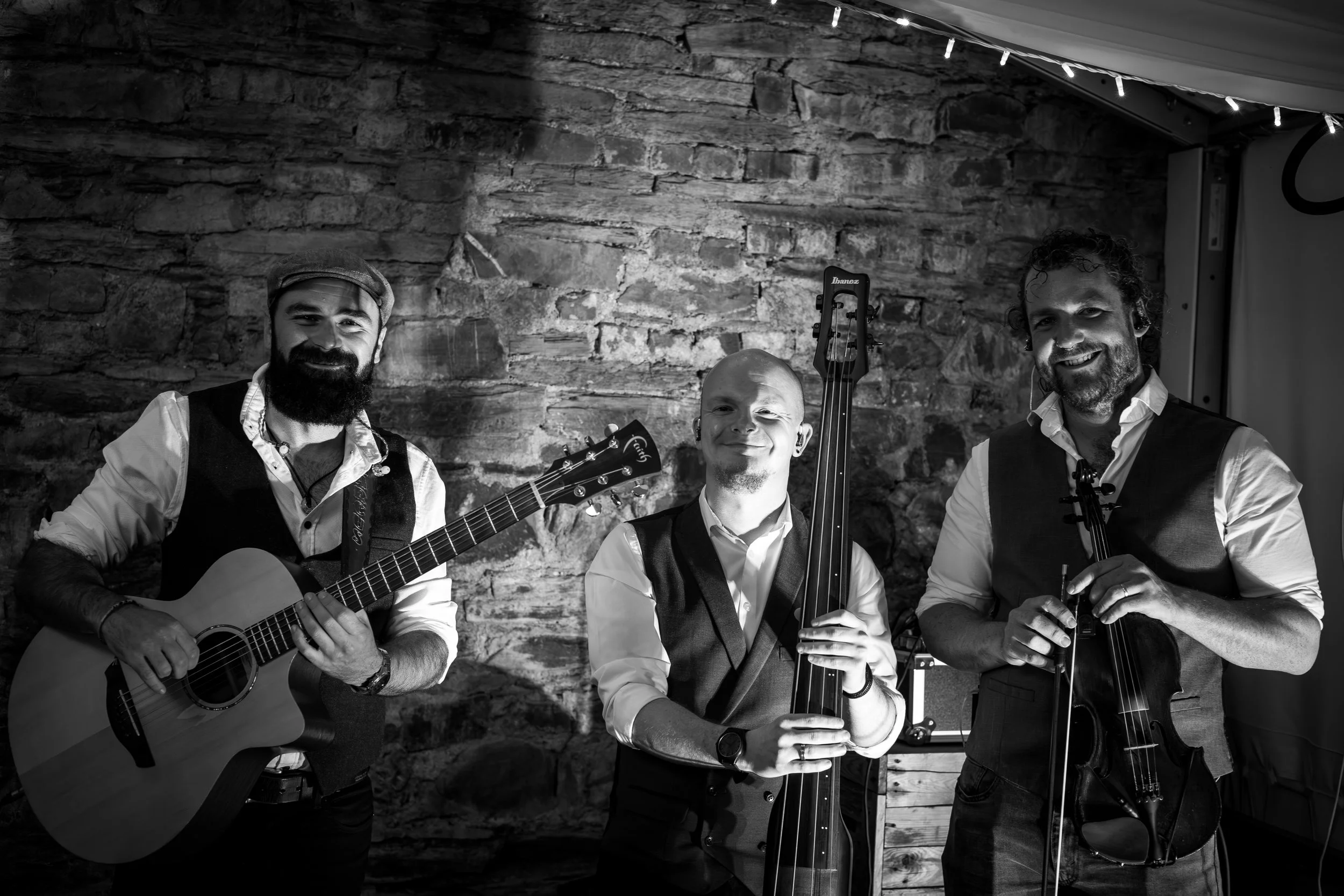 Three men, two with guitars and one with a violin, posing in front of a brick wall and string lights, smiling at the camera.