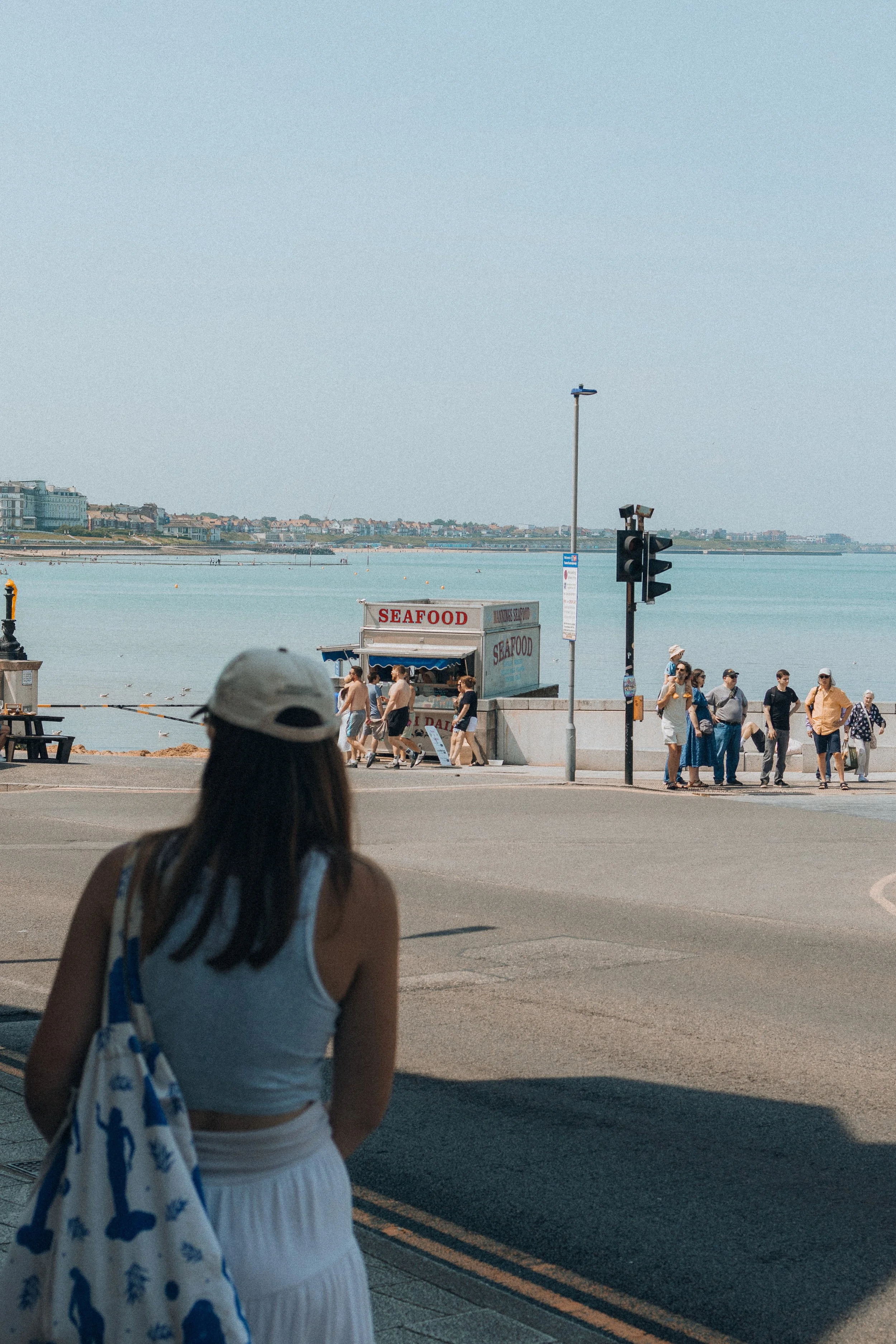 Beachside scene with people walking along the promenade, a seafood stand, and water in the background under a clear sky.