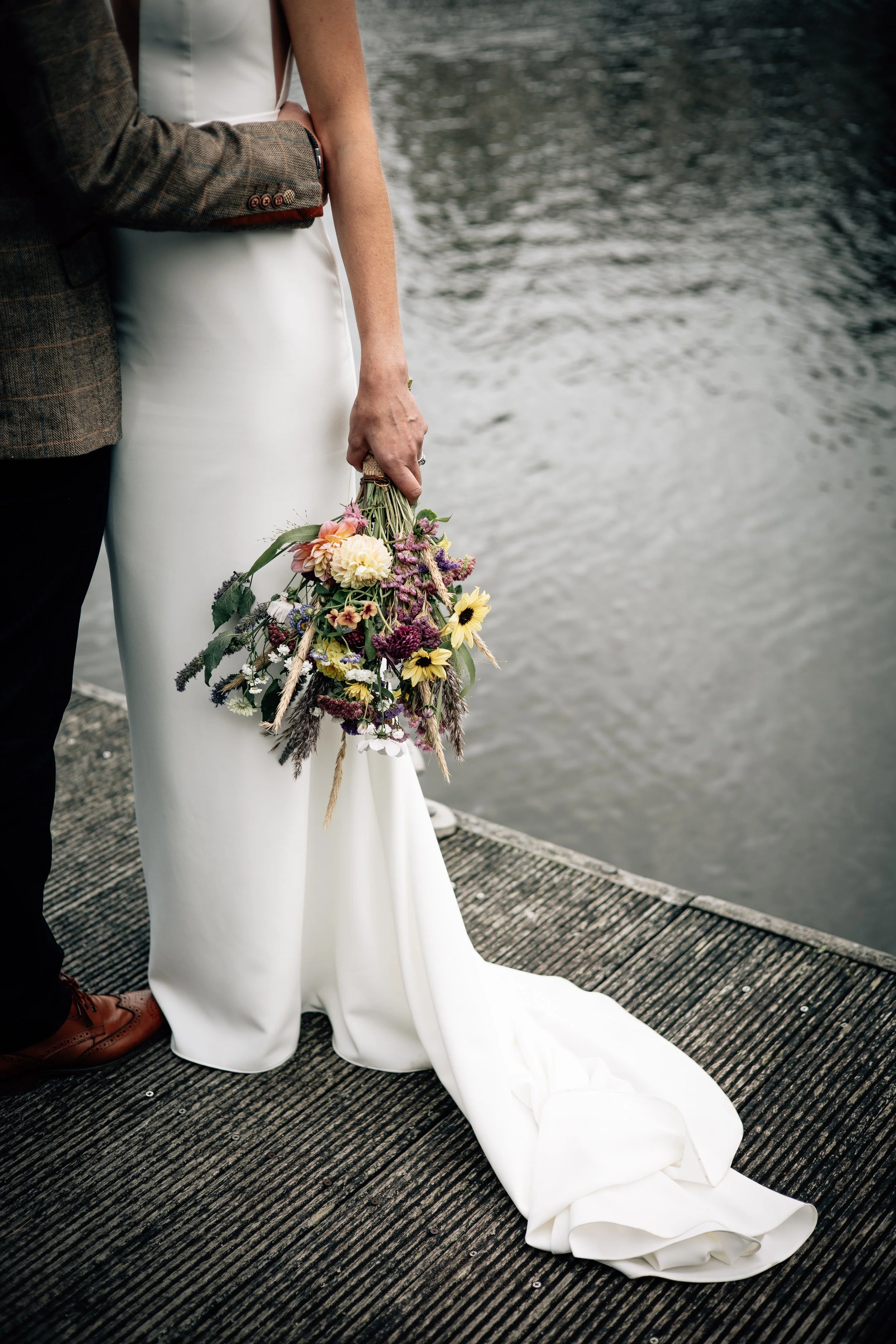 Close-up of a bride in a white wedding dress holding a bouquet of flowers, standing on a wooden dock by the water, with a groom partially visible beside her.