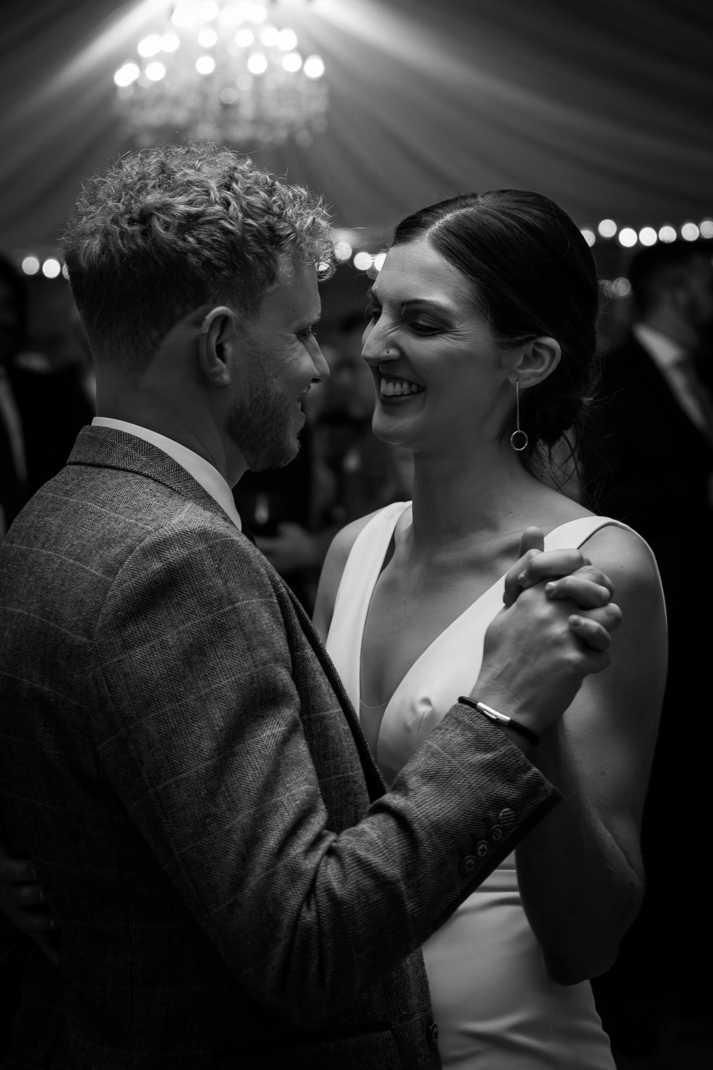 A black-and-white photo of a man and woman dancing closely, smiling at each other, in a festive setting with a chandelier and string lights in the background.