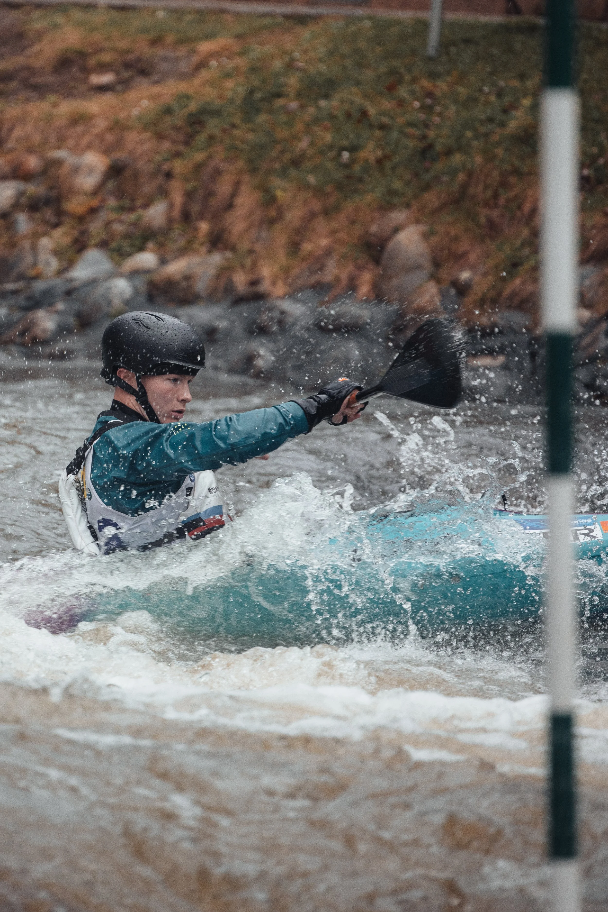 A person wearing a black helmet and blue jacket paddling a kayak in whitewater rapids, with a rocky and grassy bank in the background.