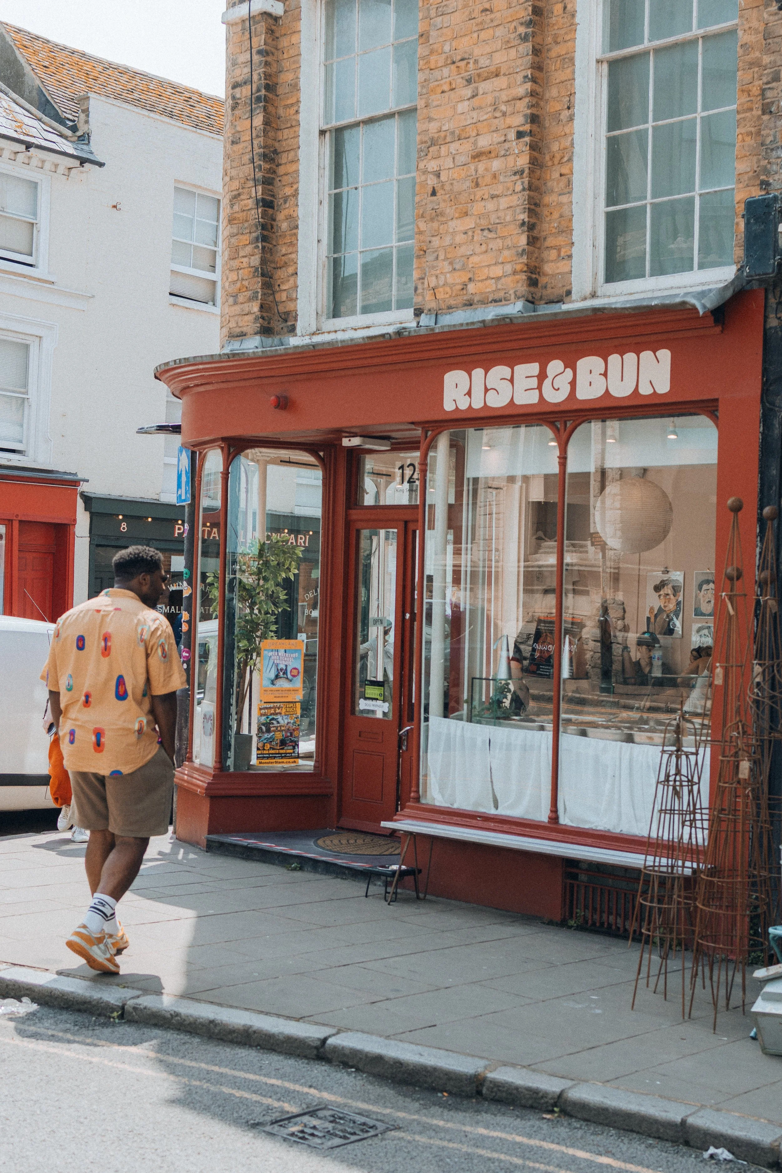 A man with curly hair wearing a beige shirt with colorful patterns, gray shorts, white socks, and orange sneakers walking on a sidewalk in front of a red storefront named "Rise & Bun" with large windows, a door, and some outdoor furniture. The buildi