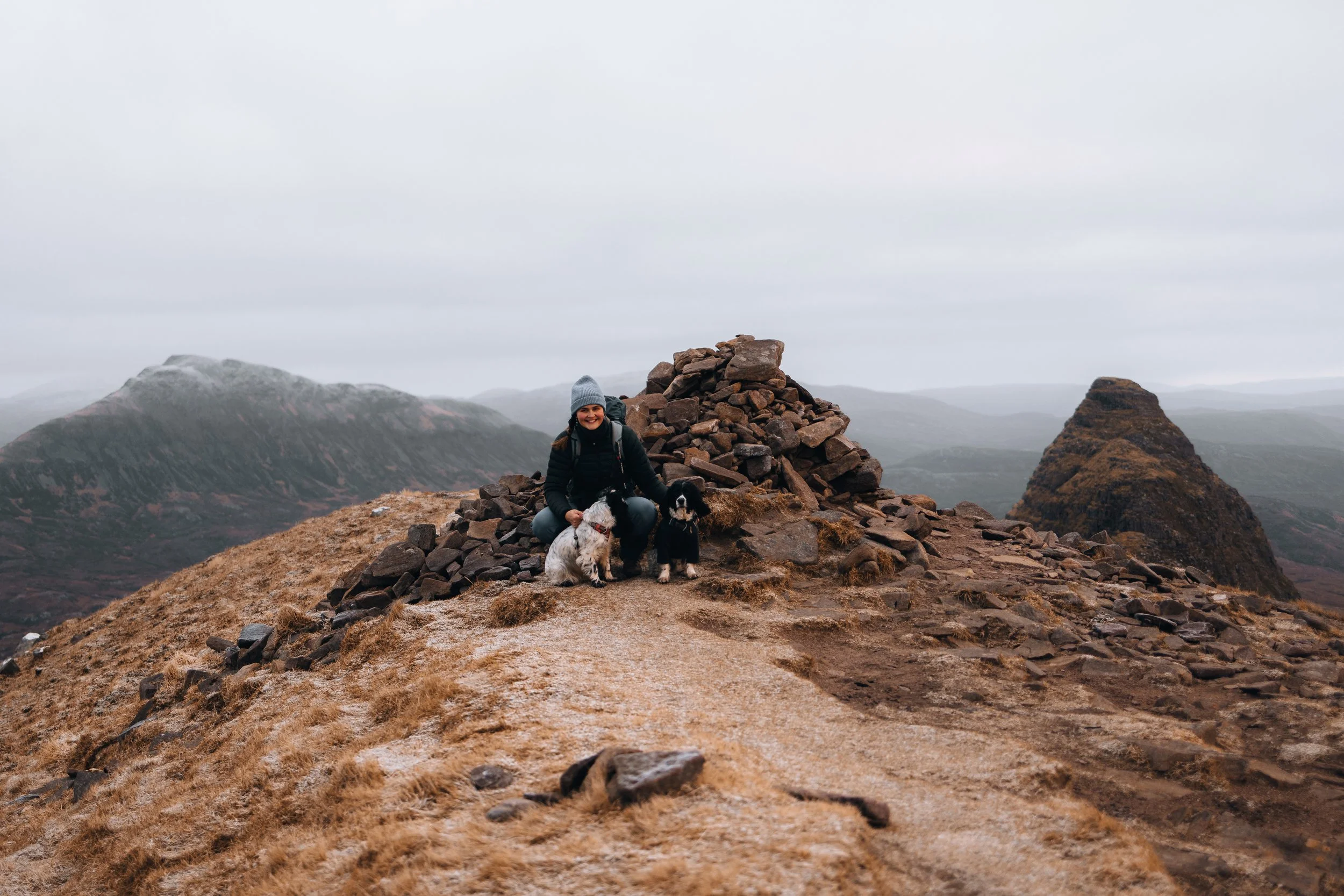 A woman in outdoor clothing and a gray beanie smiling while kneeling on a rocky mountain summit with two dogs, surrounded by a rugged mountain landscape under a cloudy sky.