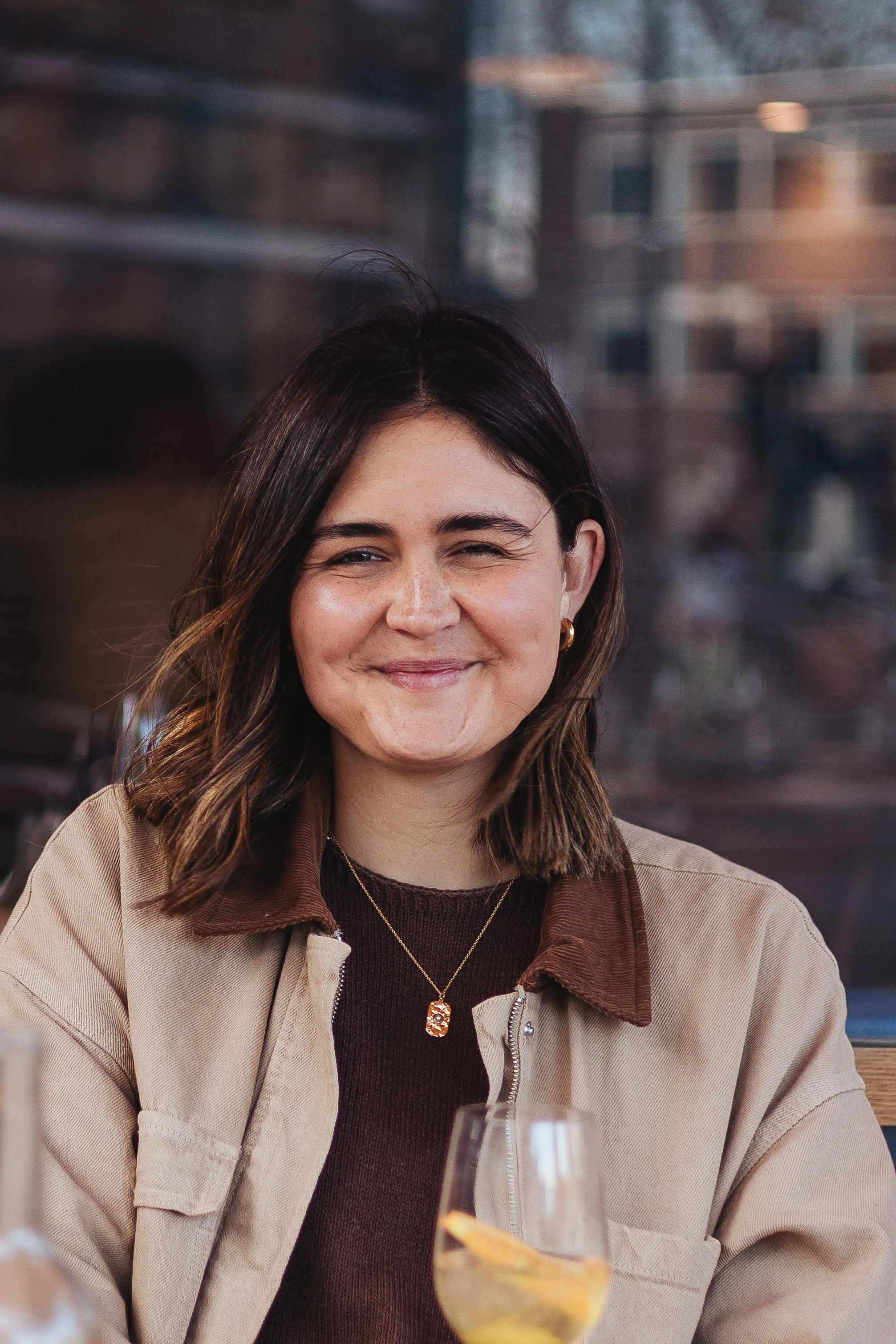 A woman smiling at an outdoor gathering, with a blurred background of a cityscape and visible power lines, dressed in a beige jacket and brown top, wearing earrings and a necklace.