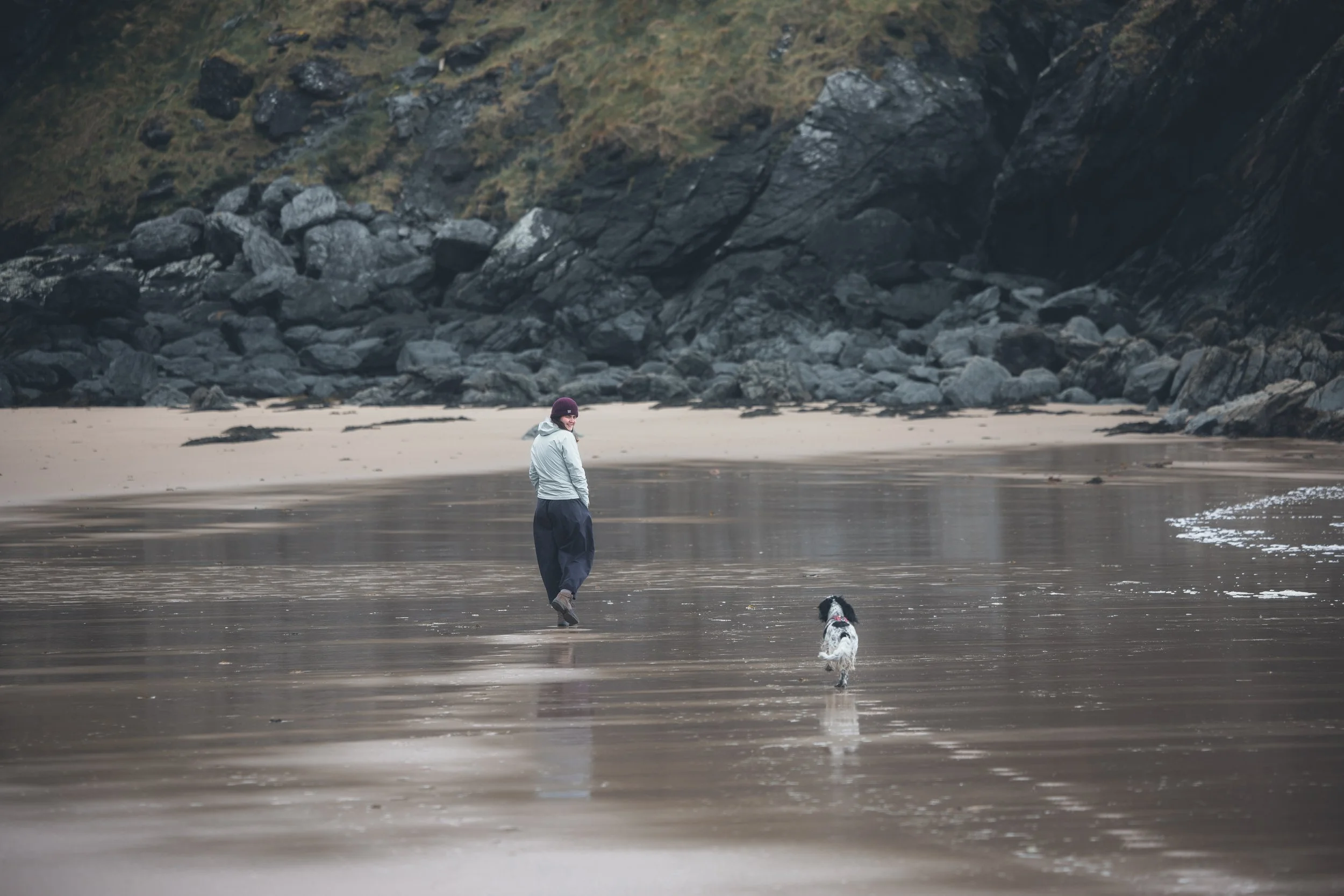 A person in a white jacket and black hat walking along the wet beach with a dog, with large dark rocks and steep hills in the background.