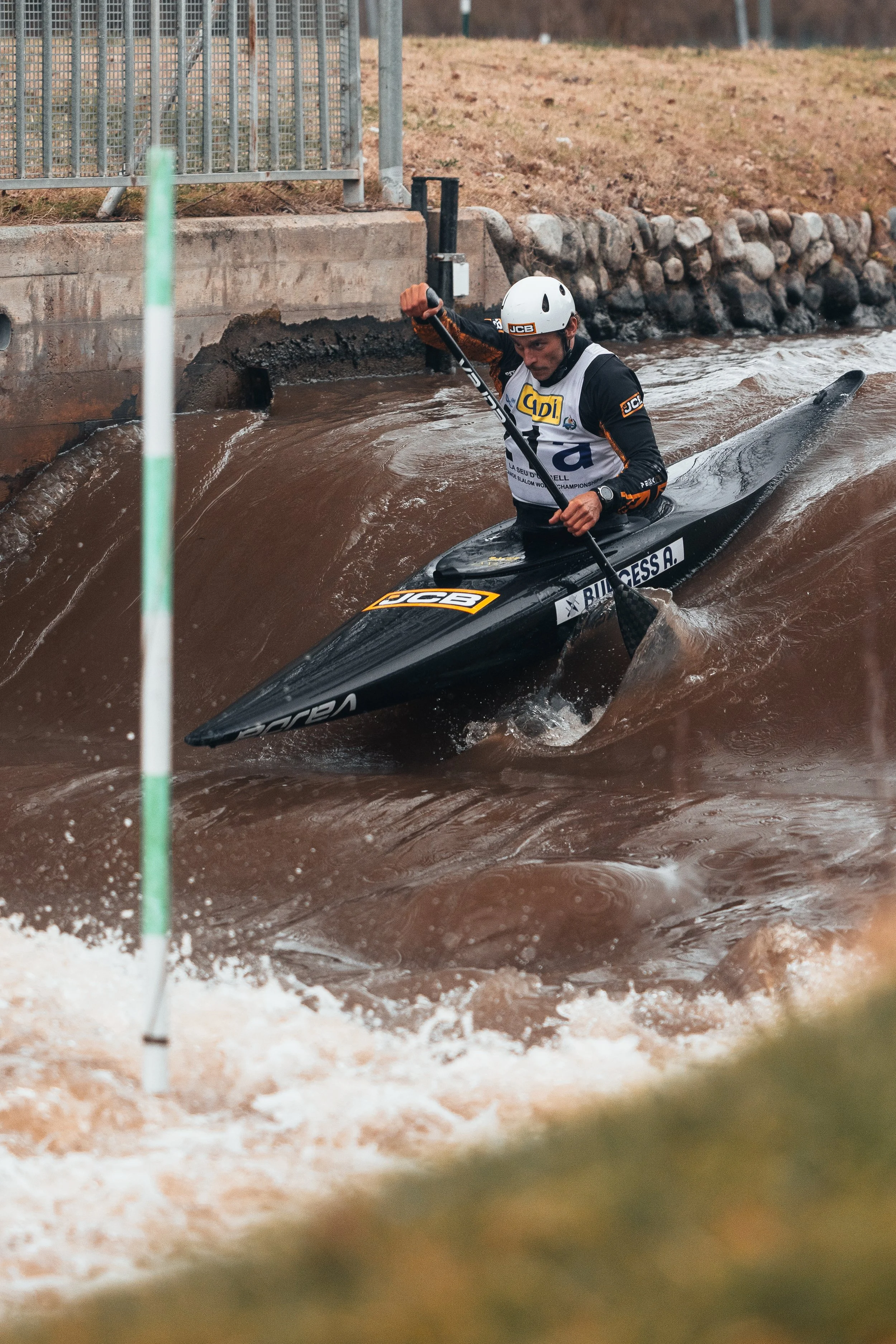 A kayaker wearing a white helmet and black and orange gear paddling through a brown water canal, approaching a gate in the water.