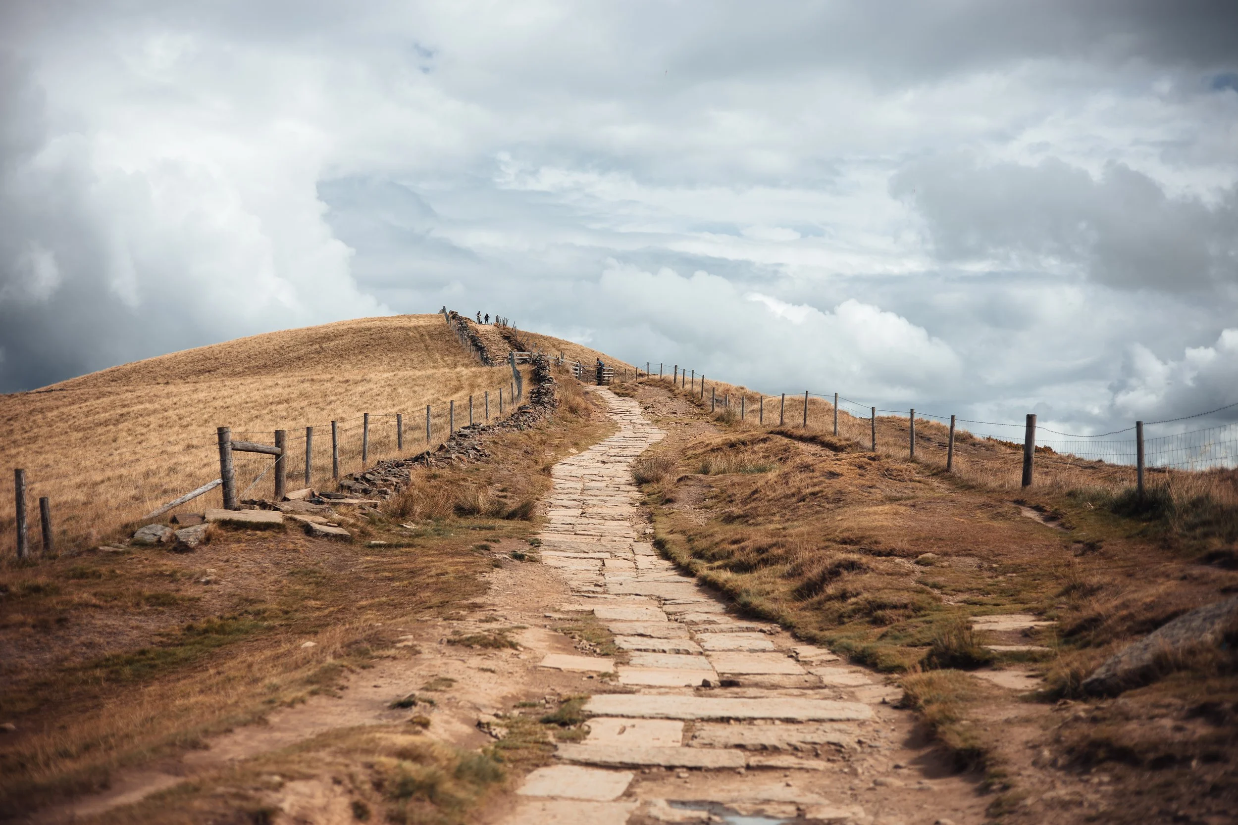 A stone-paved path winding up a hill with dry grass and a fence on each side, under a cloudy sky.