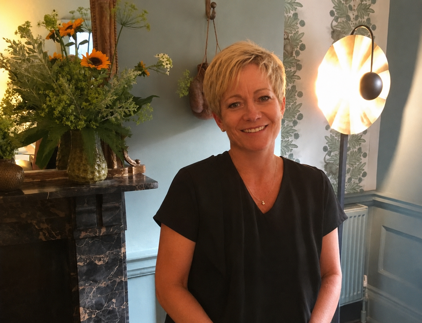 A woman with short blonde hair smiling, standing in a room with floral decor and a large bouquet of flowers on a marble fireplace mantle. Warm lighting and patterned wallpaper are visible in the background.