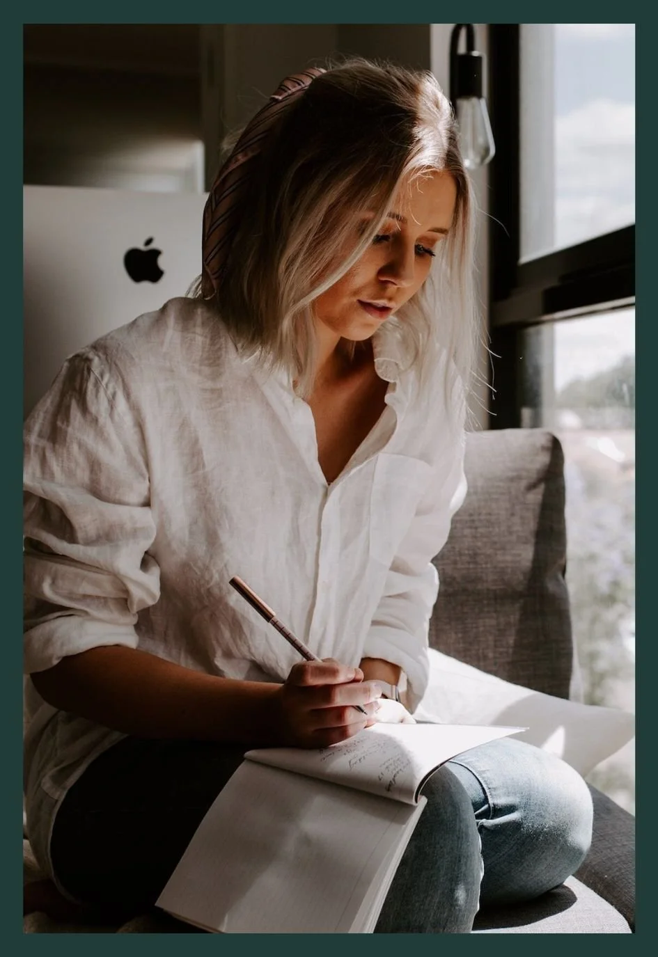 A woman with blonde hair, wearing a white blouse and a striped headband, sitting on a gray couch near a window, writing in a notebook with a pen.