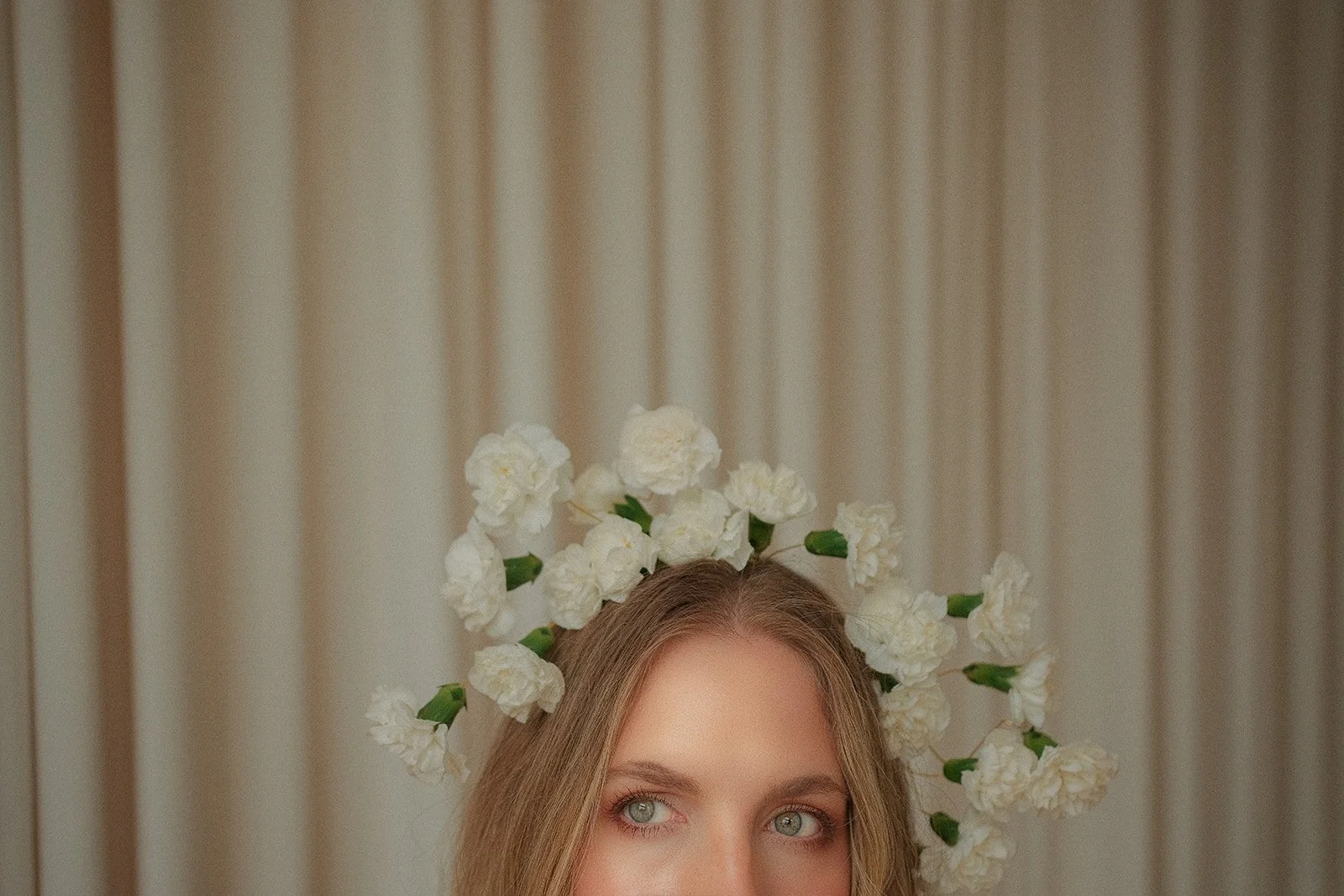 A woman with light hair and blue eyes is wearing a floral headpiece made of white flowers and green leaves, with beige curtains in the background. A non traditional bride.