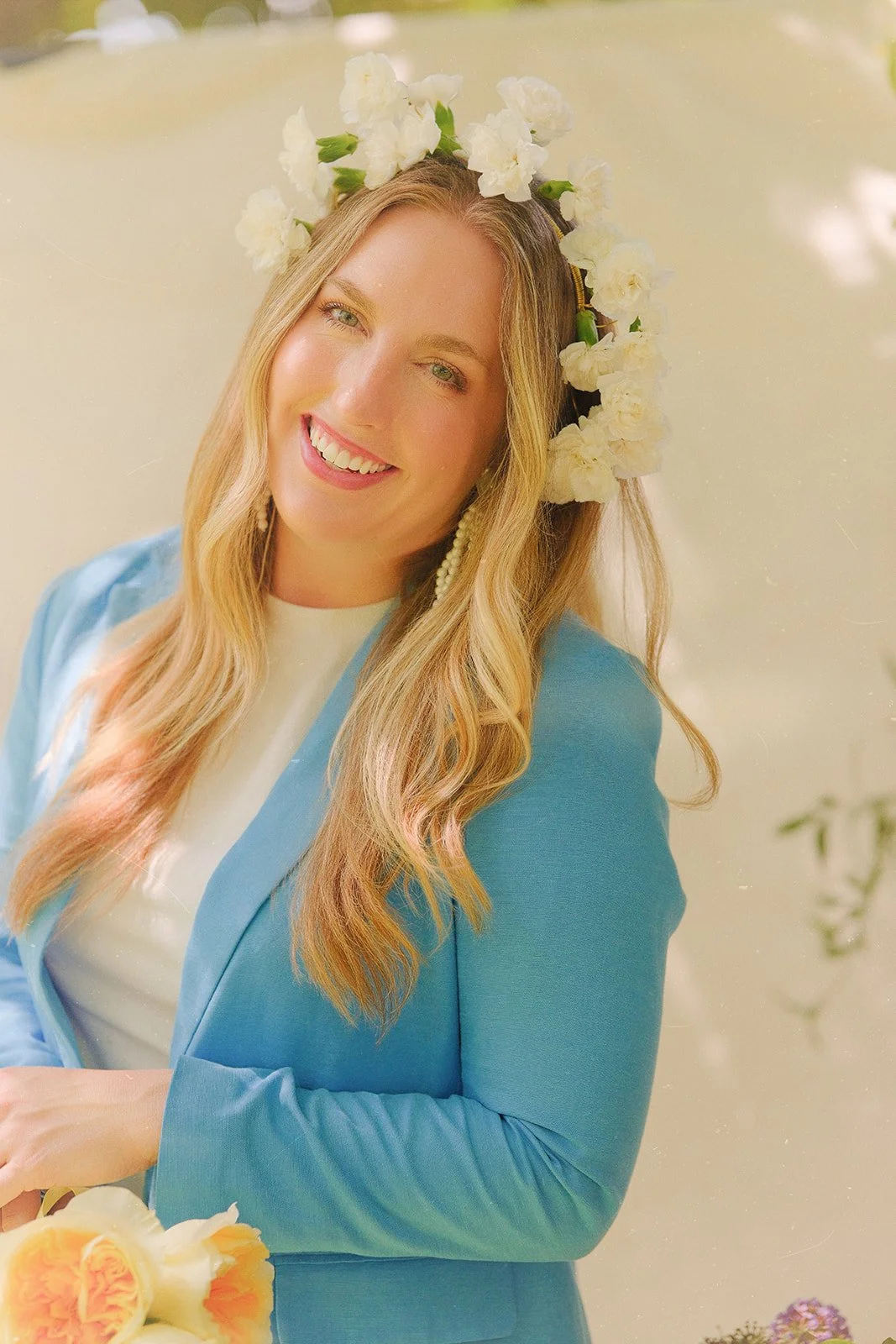 A woman with long blonde hair wearing a flower crown, smiling, dressed in a light blue blazer and white top, holding a bouquet of orange and white roses. A non traditional bride.