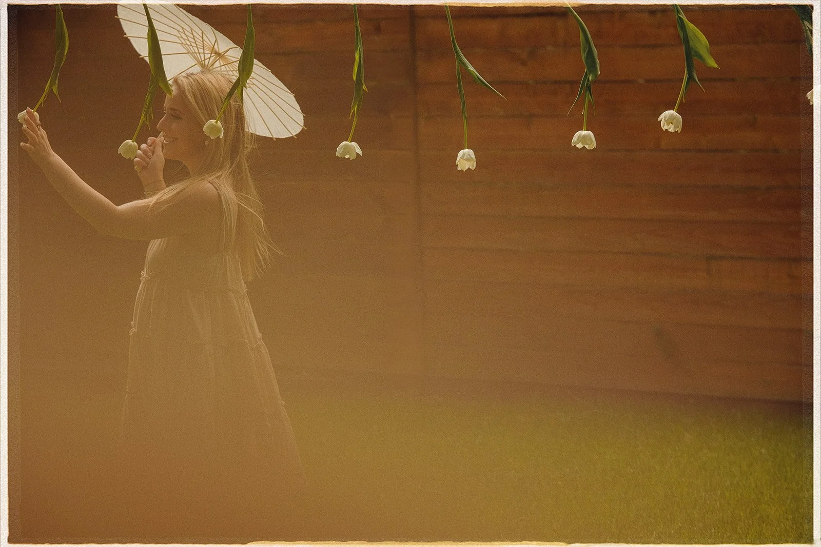 A woman in a brown dress holding a white parasol and white flowers, standing outdoors near a wooden fence. A woman playing with tulips hanging from a clothesline.