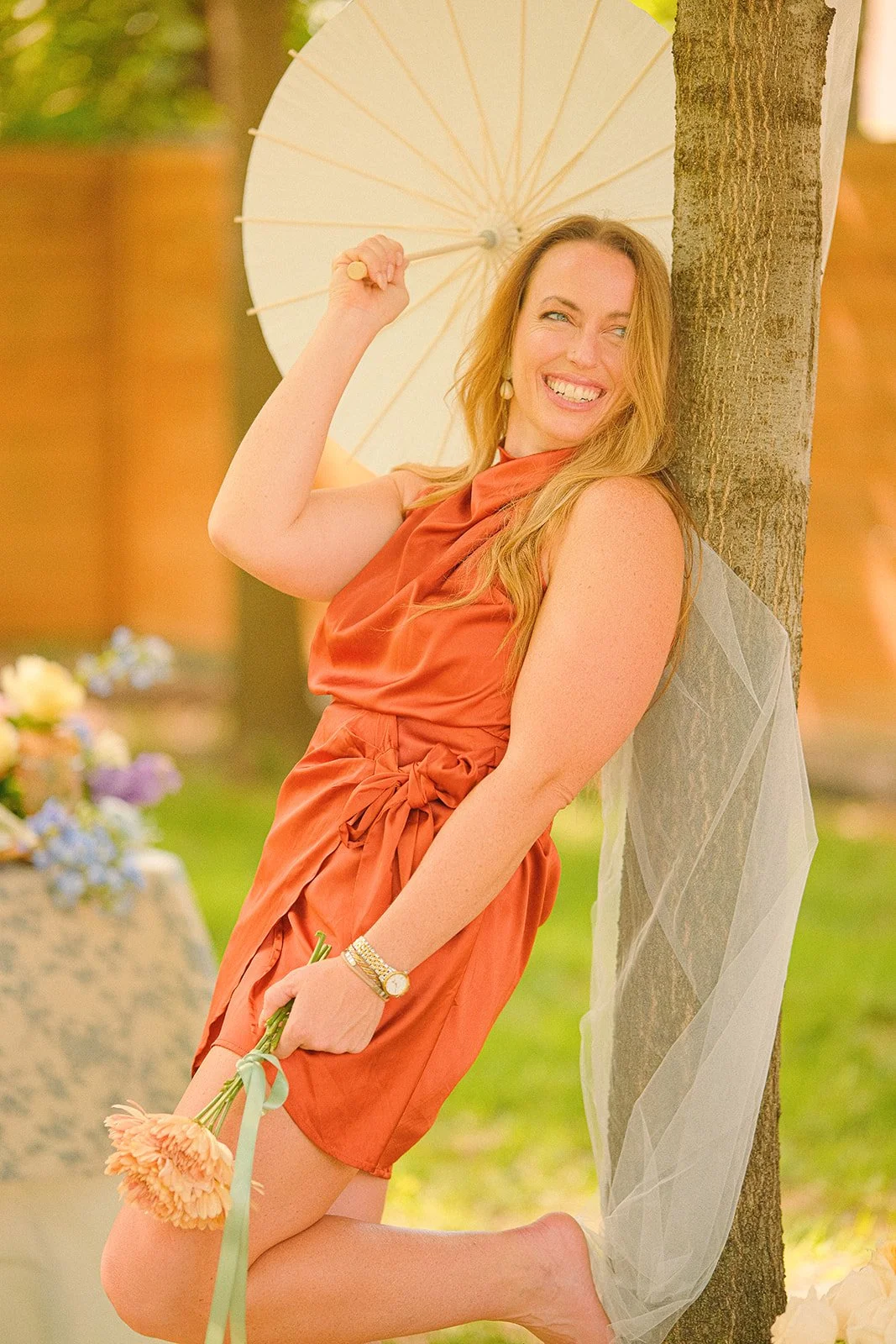 A woman kneeling outdoors, leaning against a tree with a sheer veil draped over it. She is holding a pink flower bouquet and a white parasol, wearing a rust-colored dress and smiling.