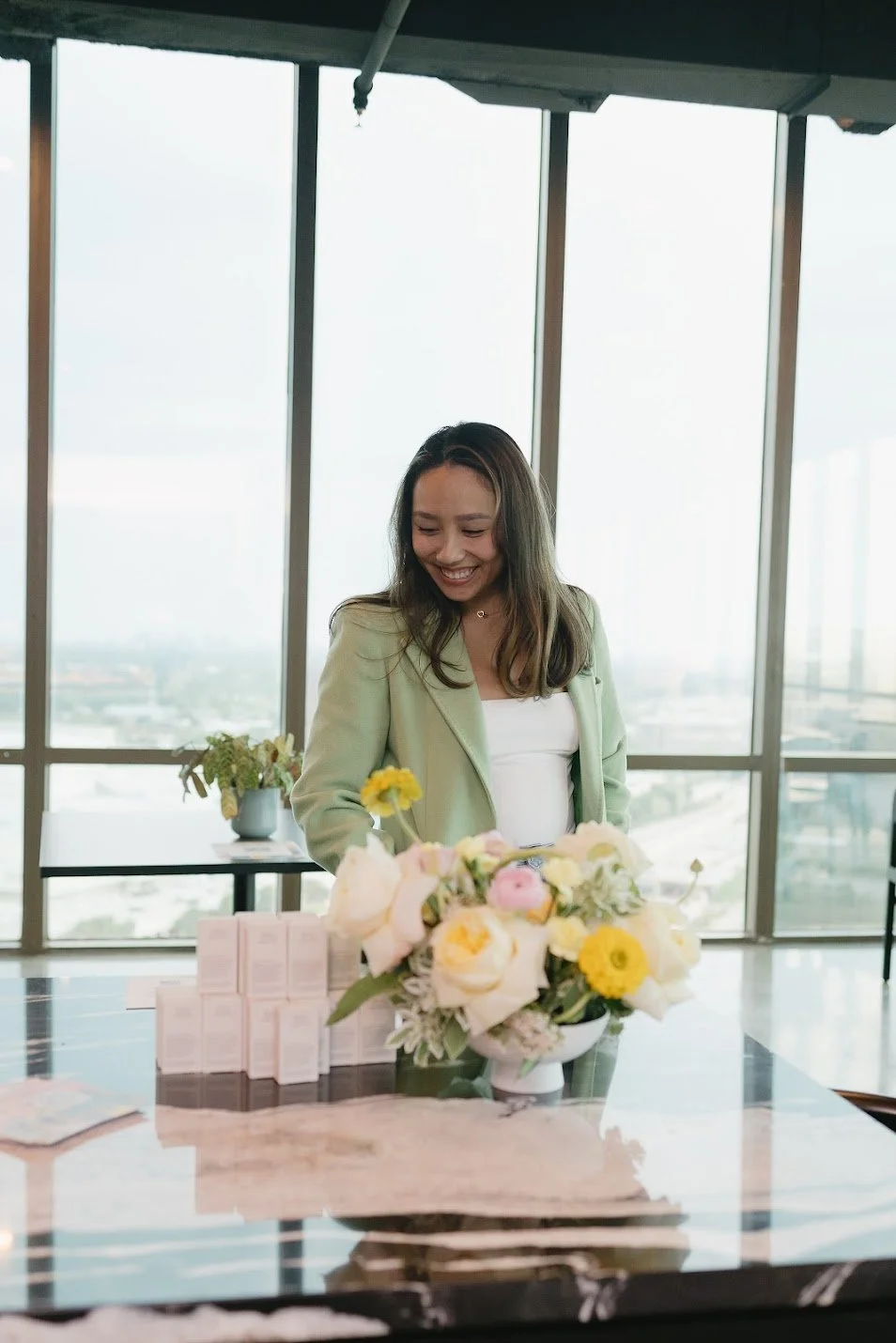 A woman in a green blazer smiling at a floral arrangement on a marble table inside a high-rise building with large windows showing a city view. A custom floral arrangement for a brand partnership event.