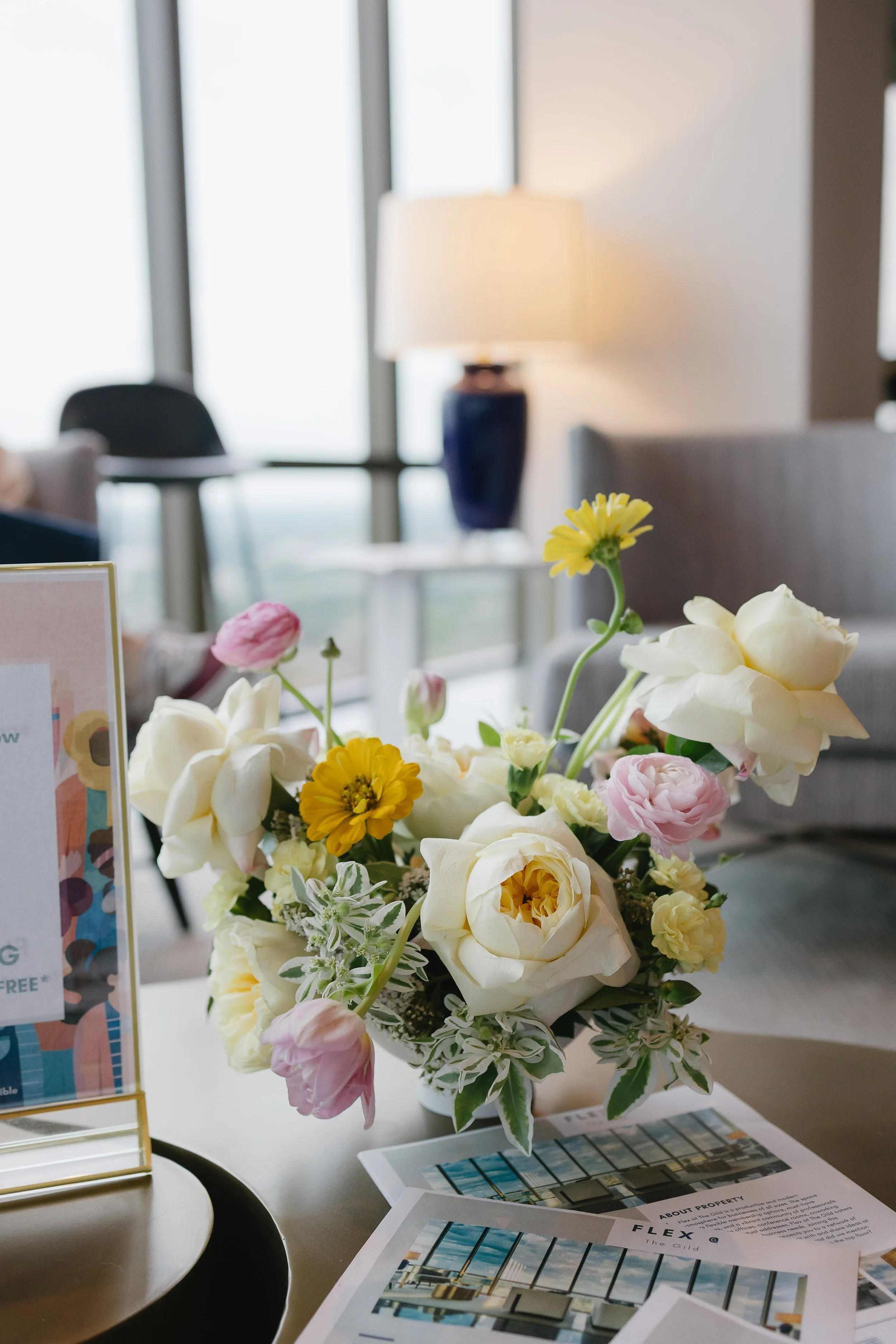 A bouquet of white, pink, and yellow flowers on a white table near some brochures and magazines, with a blurred background of a room with large windows and a popular lamp. A custom floral arrangement for a brand partnership.