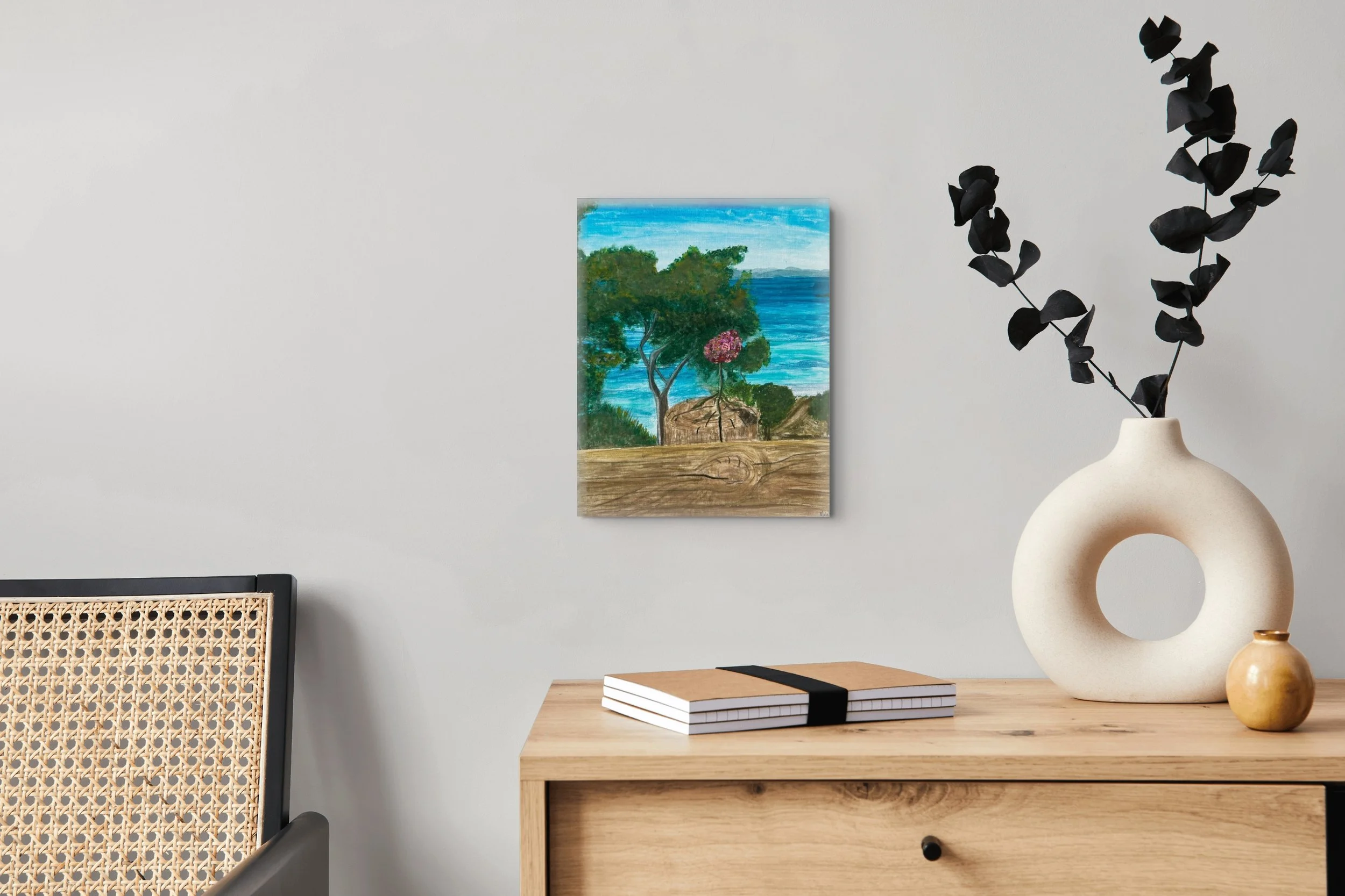 A modern interior scene with a wooden desk, a beige circular vase with dark, leafy branches, and smaller yellow and beige vases. There are stacked notebooks on the desk, a painting of a landscape with trees, a lake, and houses on the wall, and part of a woven chair is visible. The background wall is light-colored.