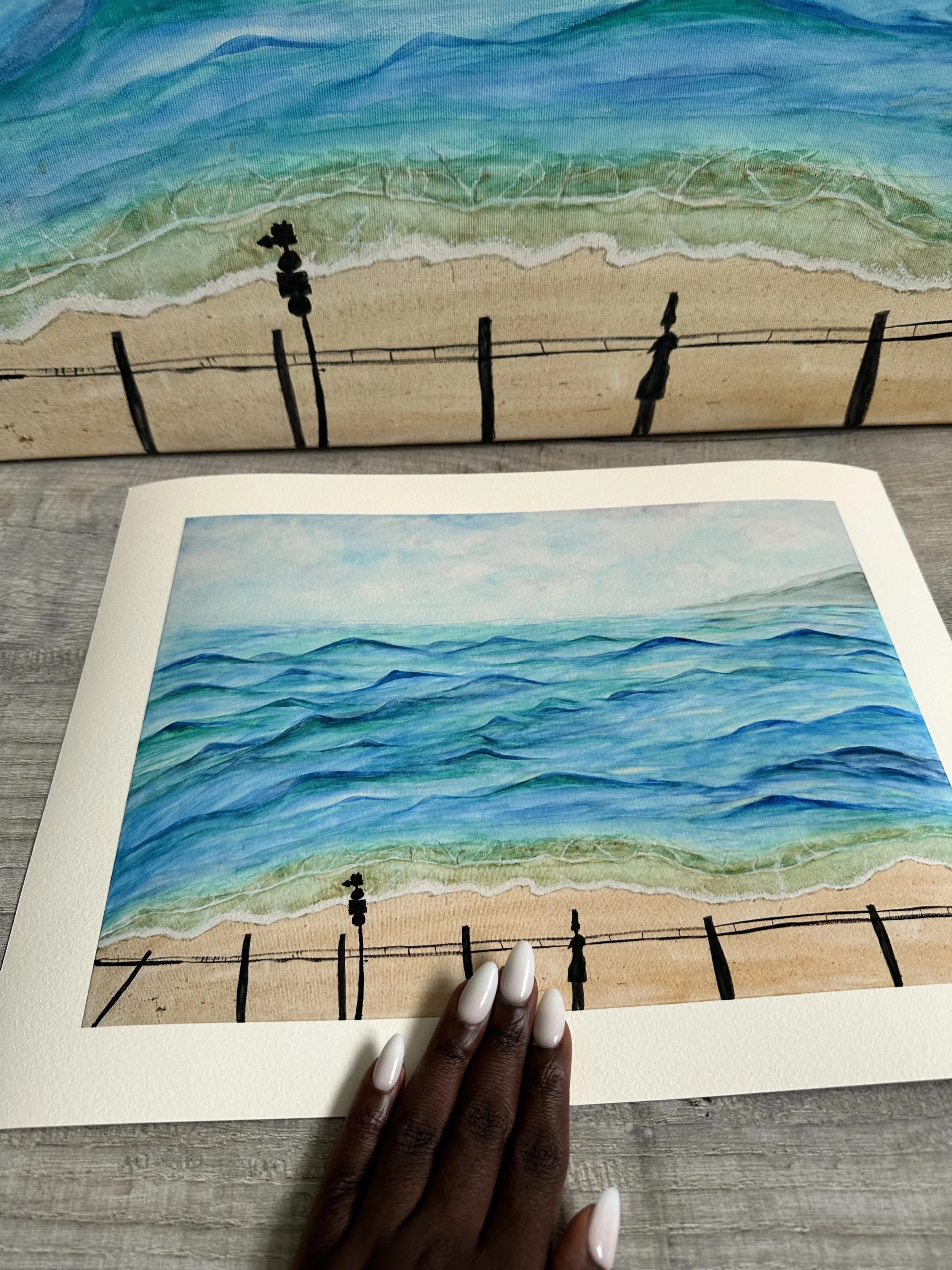 Hand holding a museum-quality giclée fine art print on Hahnemühle William Turner 310 gsm, a 100% cotton, of a watercolor painting of a beach scene with blue ocean waves, sandy shore, and a fence with posts and wire, viewed from above.
