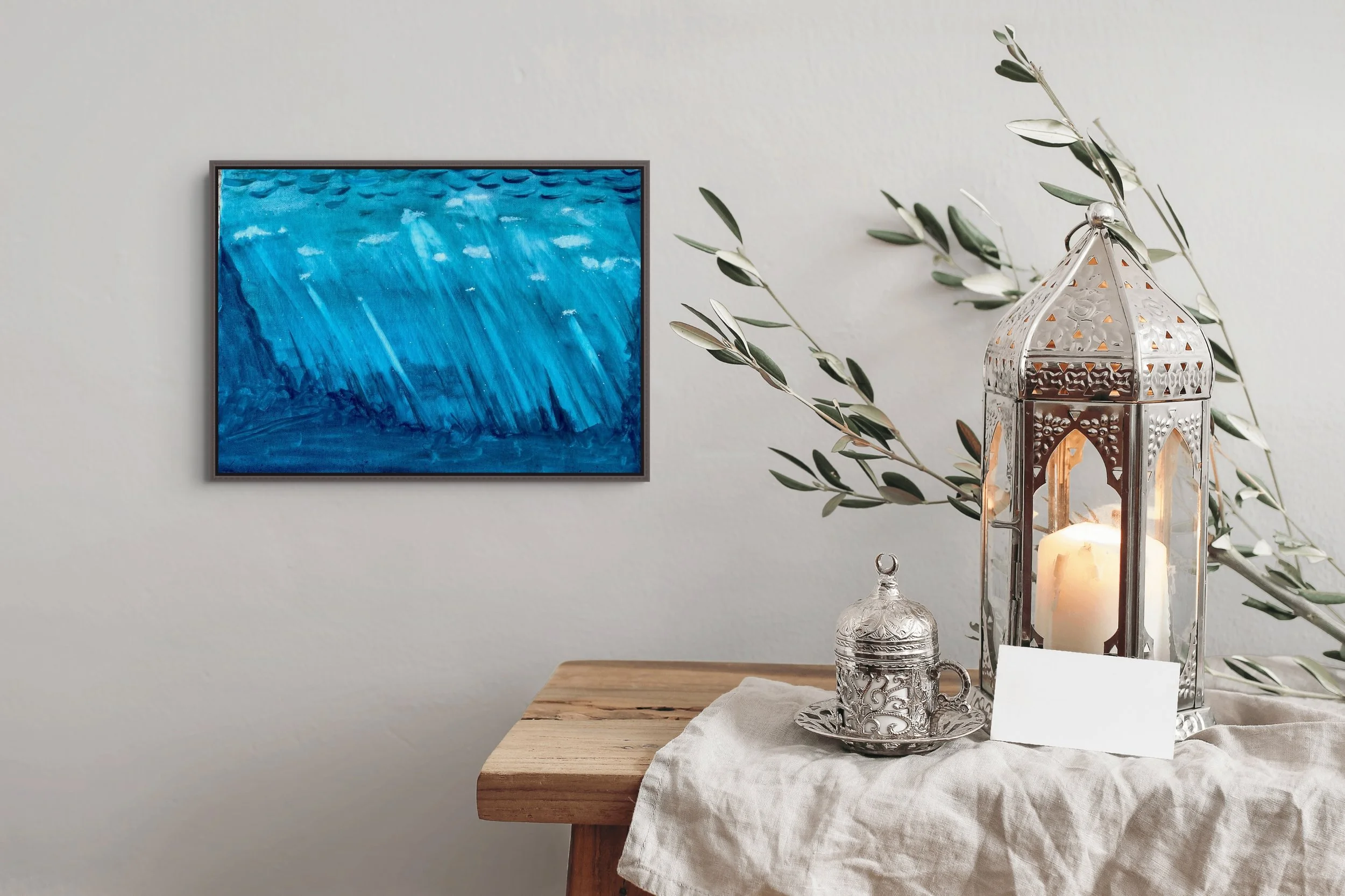A decorative table setting with a silver lantern, a candle inside, a silver ornate cup, and a leafy branch on a beige tablecloth, against a white wall with a blue abstract painting.