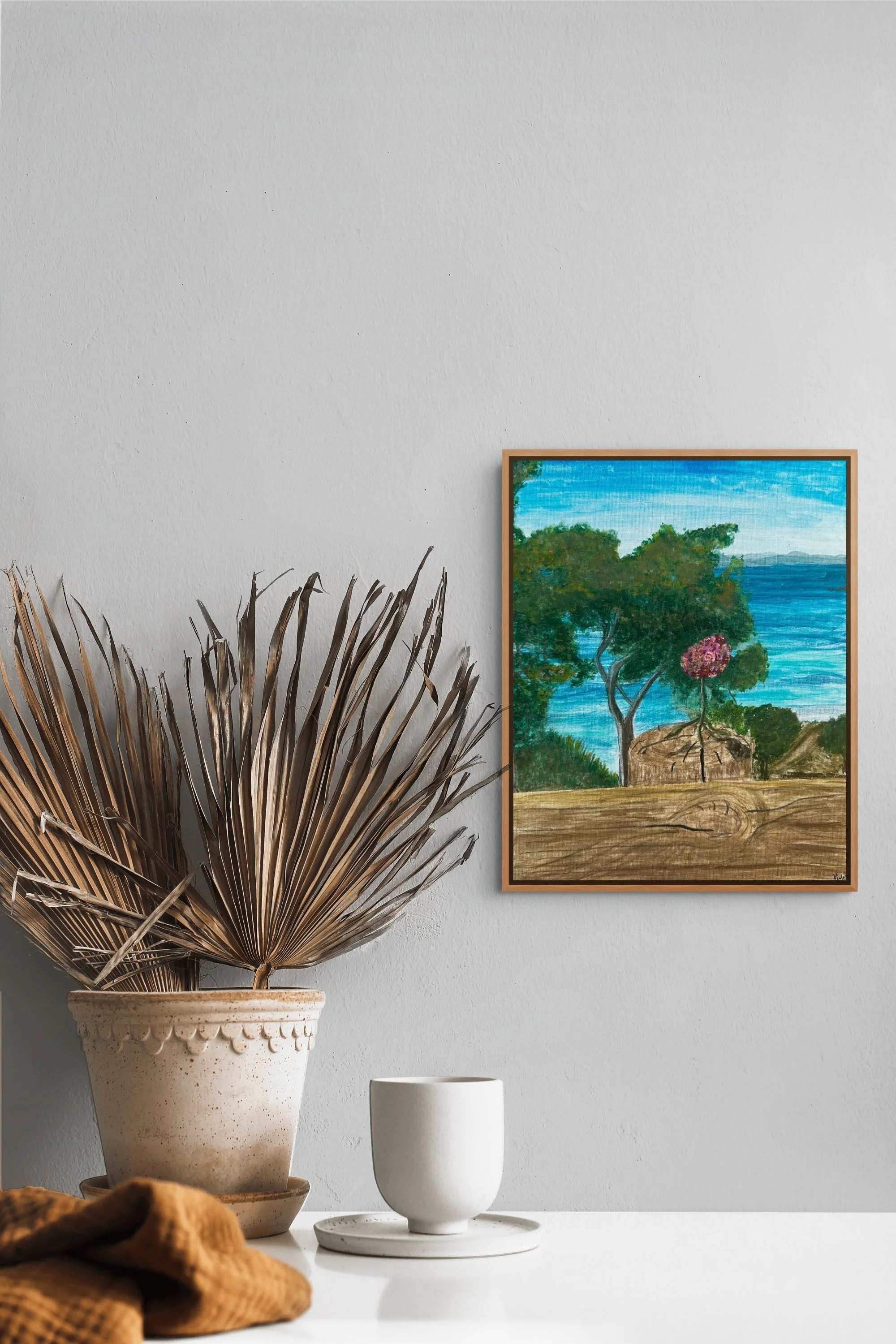 Interior decor scene featuring a large dried palm leaf in a decorative ceramic pot on a white table, with two white cups and a cloth in the foreground. A framed landscape painting showing trees and water hangs on a white wall behind.