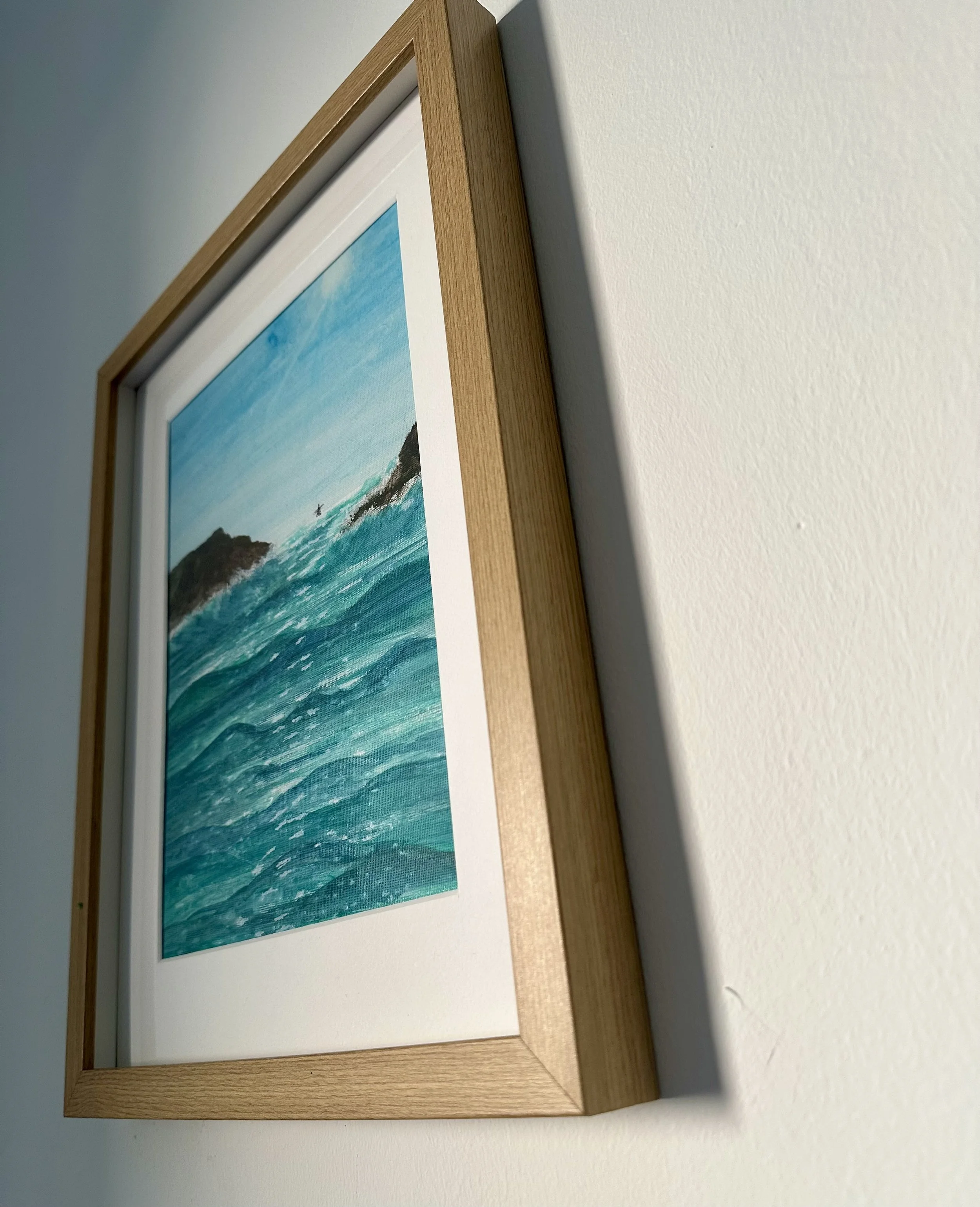 Framed watercolor painting of ocean waves viewed from a side angle, hanging on a white wall.