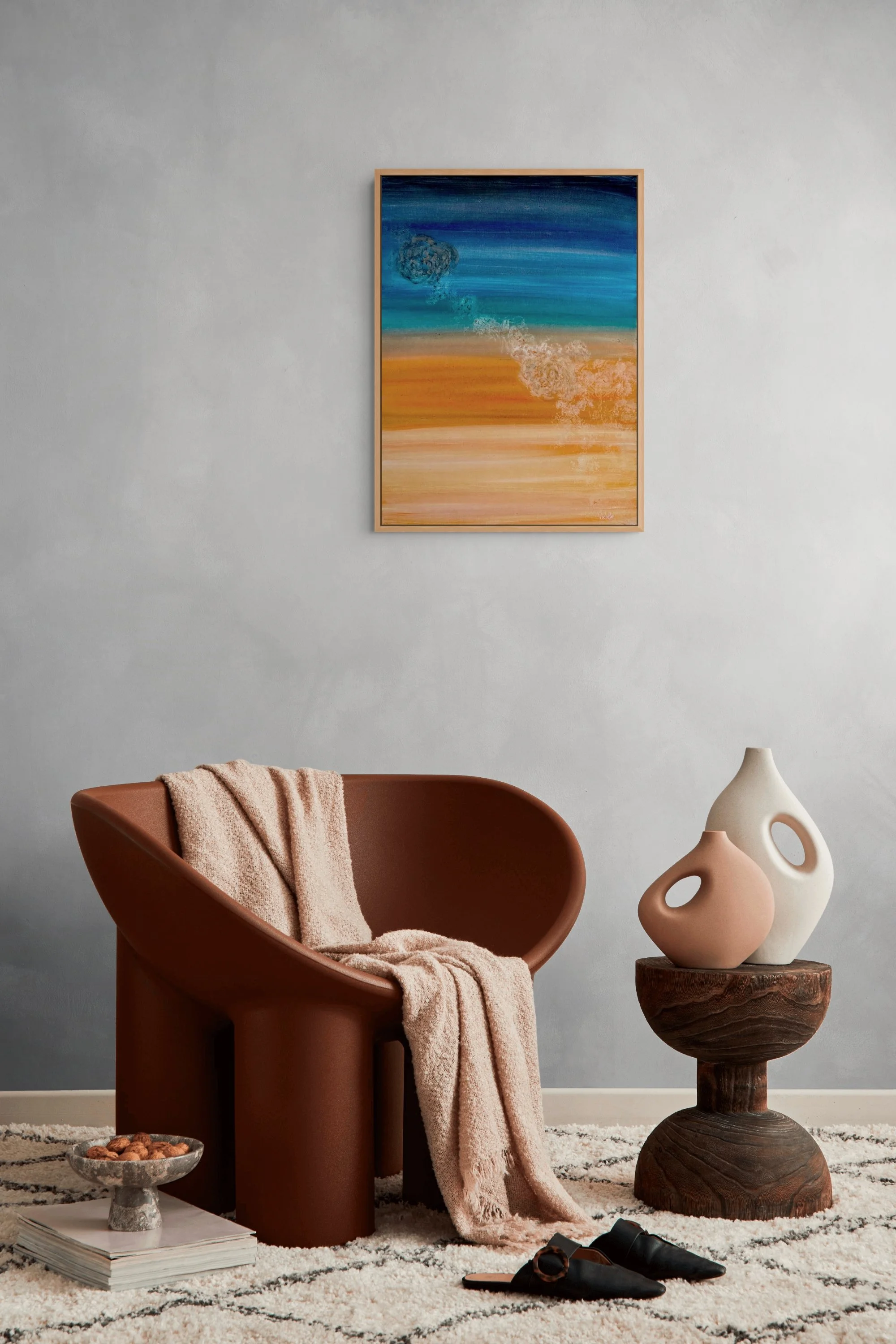 Interior of a modern, minimal living space with a brown armchair, a pink plush blanket, a decorative stone bowl filled with nuts on a stack of magazines, a pair of black shoes on a rug, a wooden side table with abstract ceramic vases, a colorful abstract painting on a gray wall, and a beige and white patterned rug.