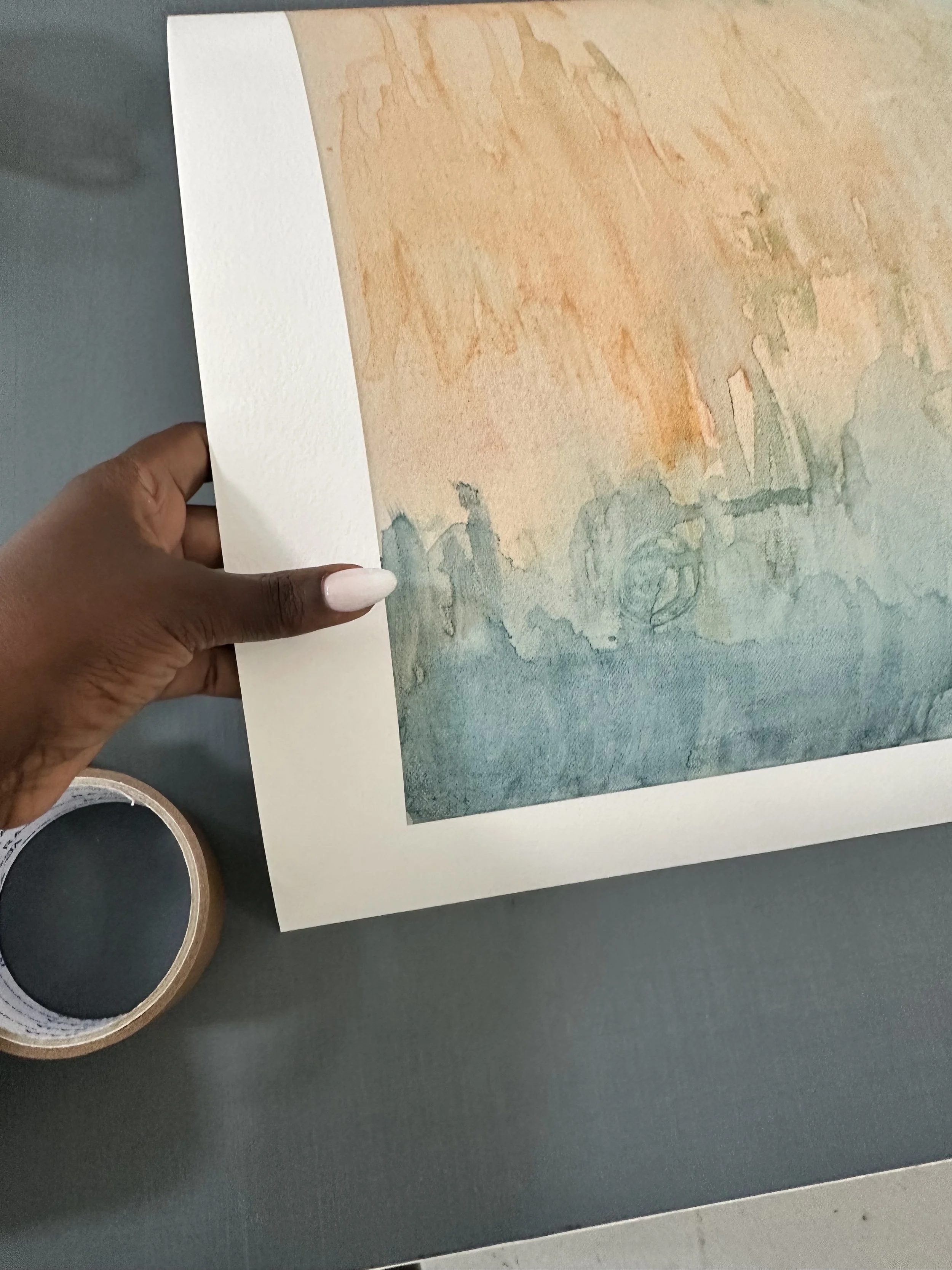 A person's hand holding a museum-quality giclée fine art print on Hahnemühle William Turner 310 gsm, a 100% cotton, of an abstract watercolor painting in shades of beige, orange, and green, with a roll of masking tape on a dark gray table.