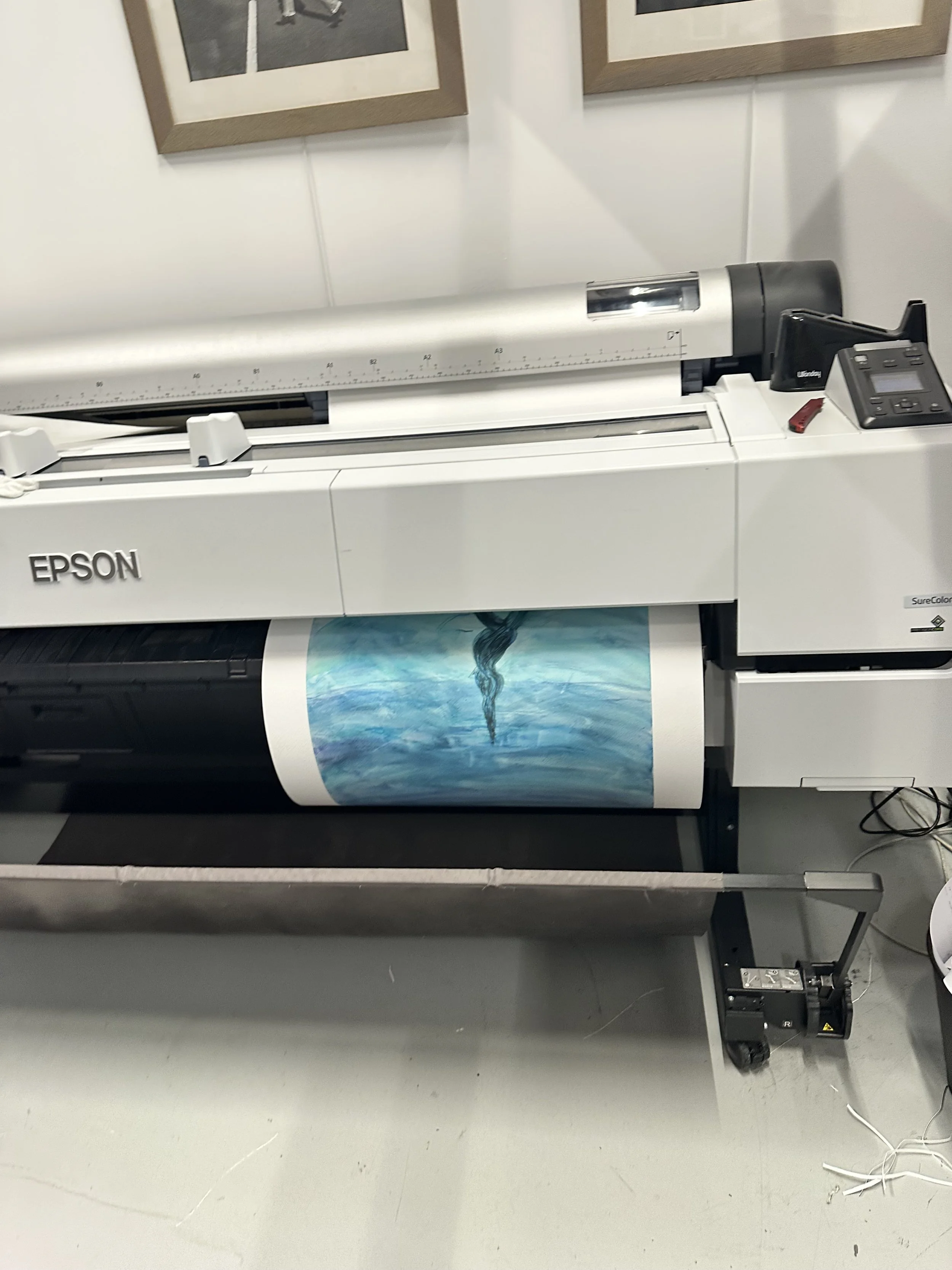 A large flatbed scanner with a fine art print of the watercolor painting, The Alchemy of Creation. The scanner is white and has an Epson logo on the front. In the background, there are two framed photographs hanging on the wall.