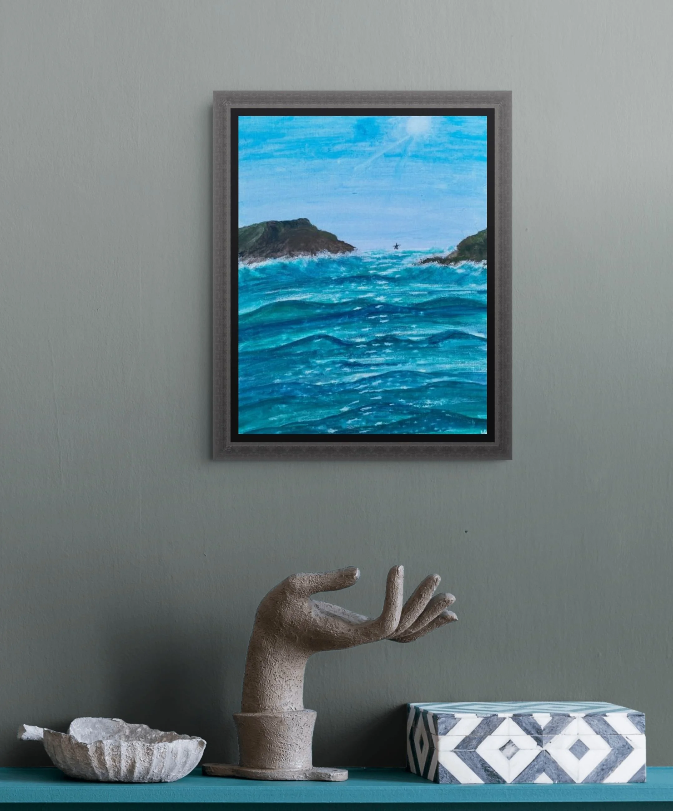 Decorative objects on a teal shelf including a textured white stone bowl, a stone urn with a large artificial white tree with thick textured branches, a framed painting of the ocean with rocks and a starfish, and a geometric blue and white box.