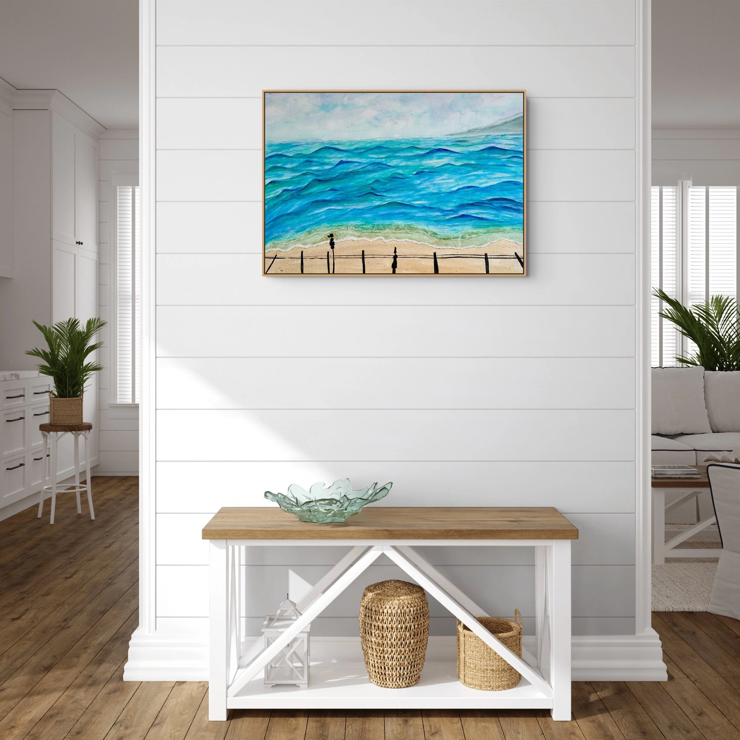 Interior of a house with a white wooden partition and a framed ocean landscape painting on the wall above a wooden table with glass decorative bowl, wicker baskets and potted plants.