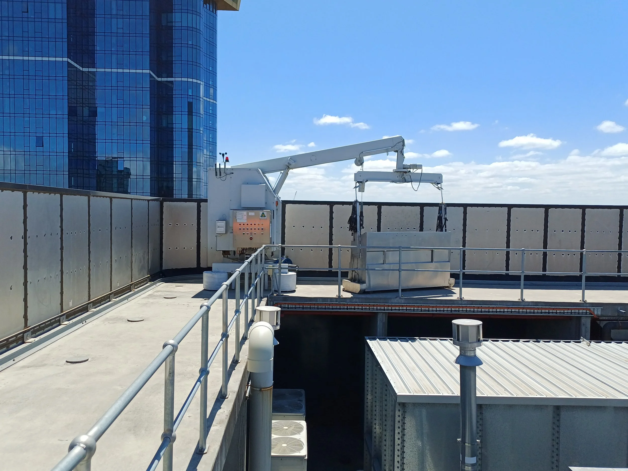 View of a rooftop with HVAC equipment, air vents, and safety railings against a backdrop of a modern glass office building and blue sky with clouds.
