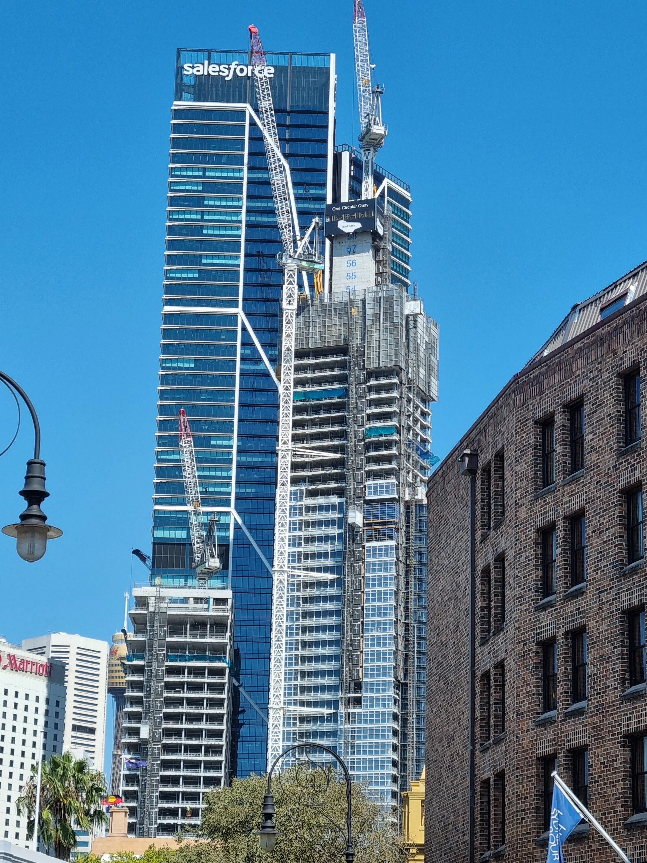 High-rise construction building with cranes, modern glass facade, and the Salesforce logo at the top, located in an urban area with other tall buildings, streetlights, and trees.