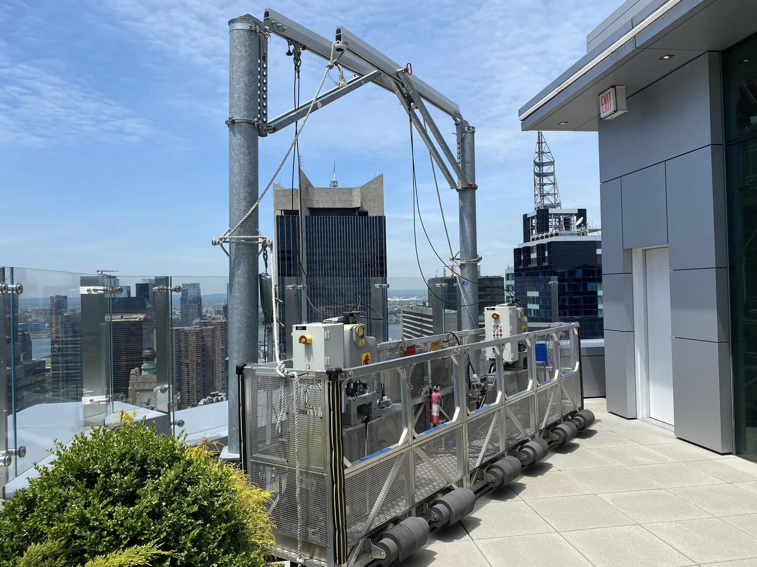 Rooftop view of a city with tall buildings including a structure with black glass windows, an antenna, and a modern building with an exit sign. The foreground shows rooftop equipment, a fire extinguisher, and lush green plants.