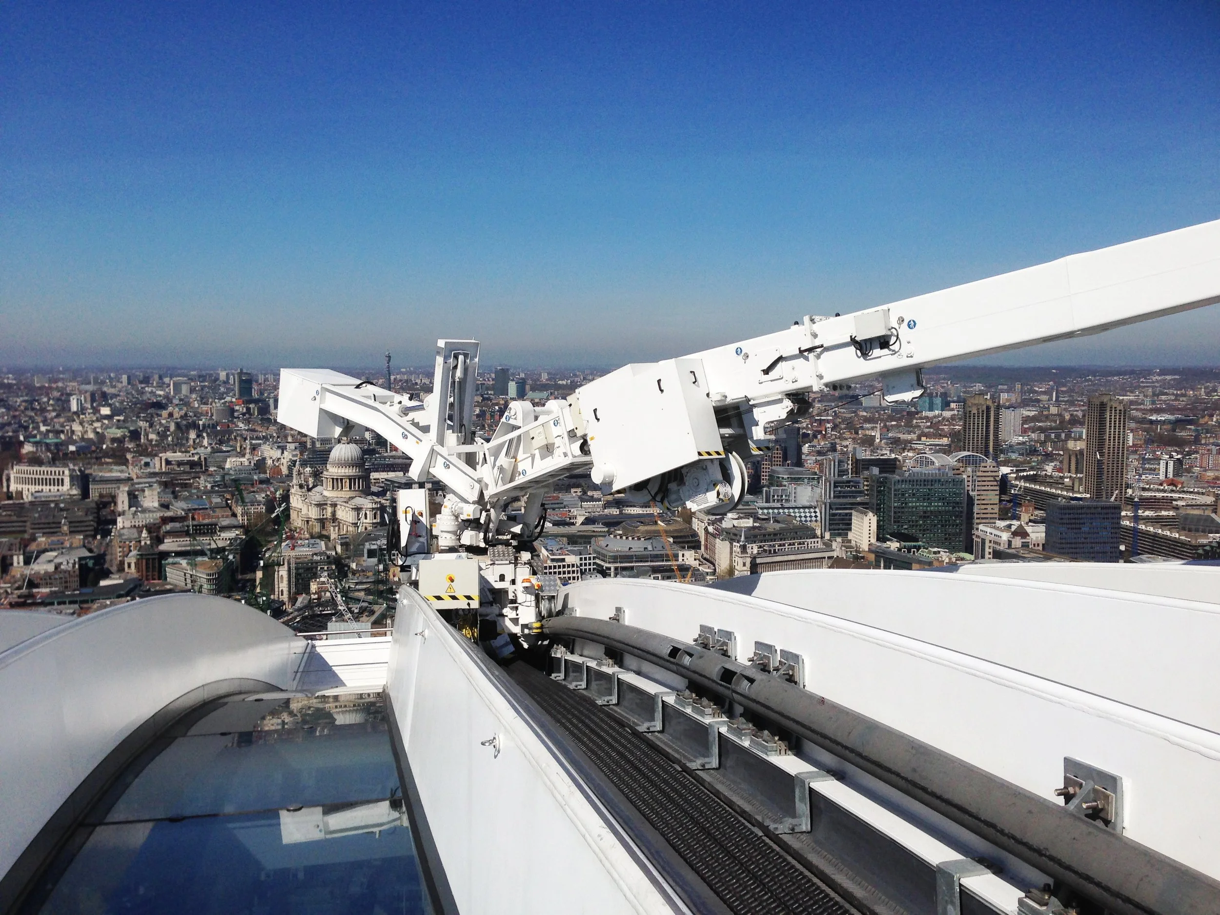 A large white construction crane perched high above a city skyline under a clear blue sky.