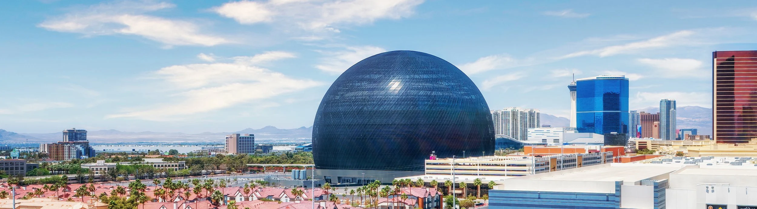 A city skyline featuring the spherical Underground Museum in Las Vegas, surrounded by various tall modern buildings, with blue skies and clouds overhead.