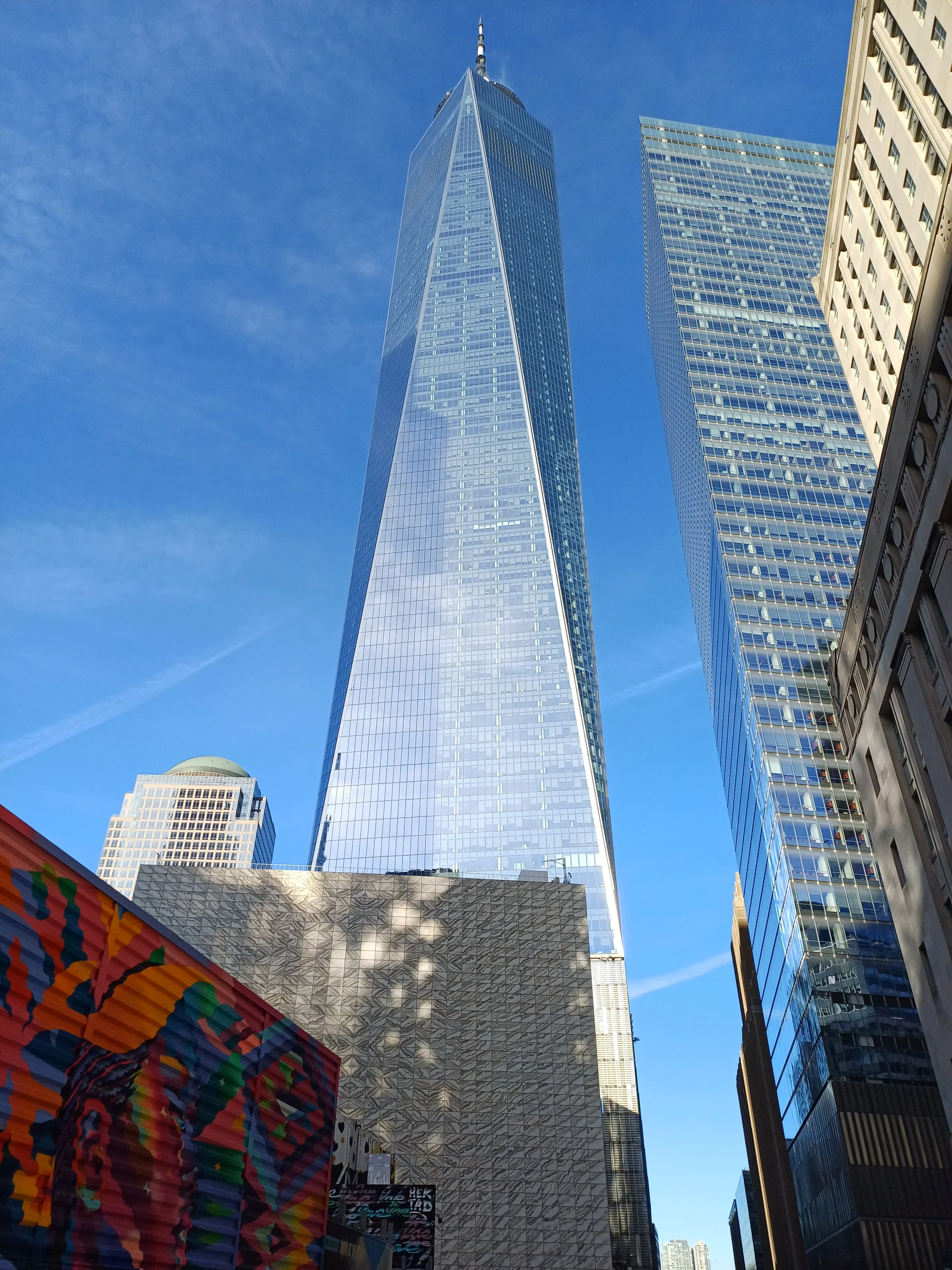 A view of One World Trade Center in New York City, with surrounding skyscrapers against a blue sky and lower colorful building in the foreground.