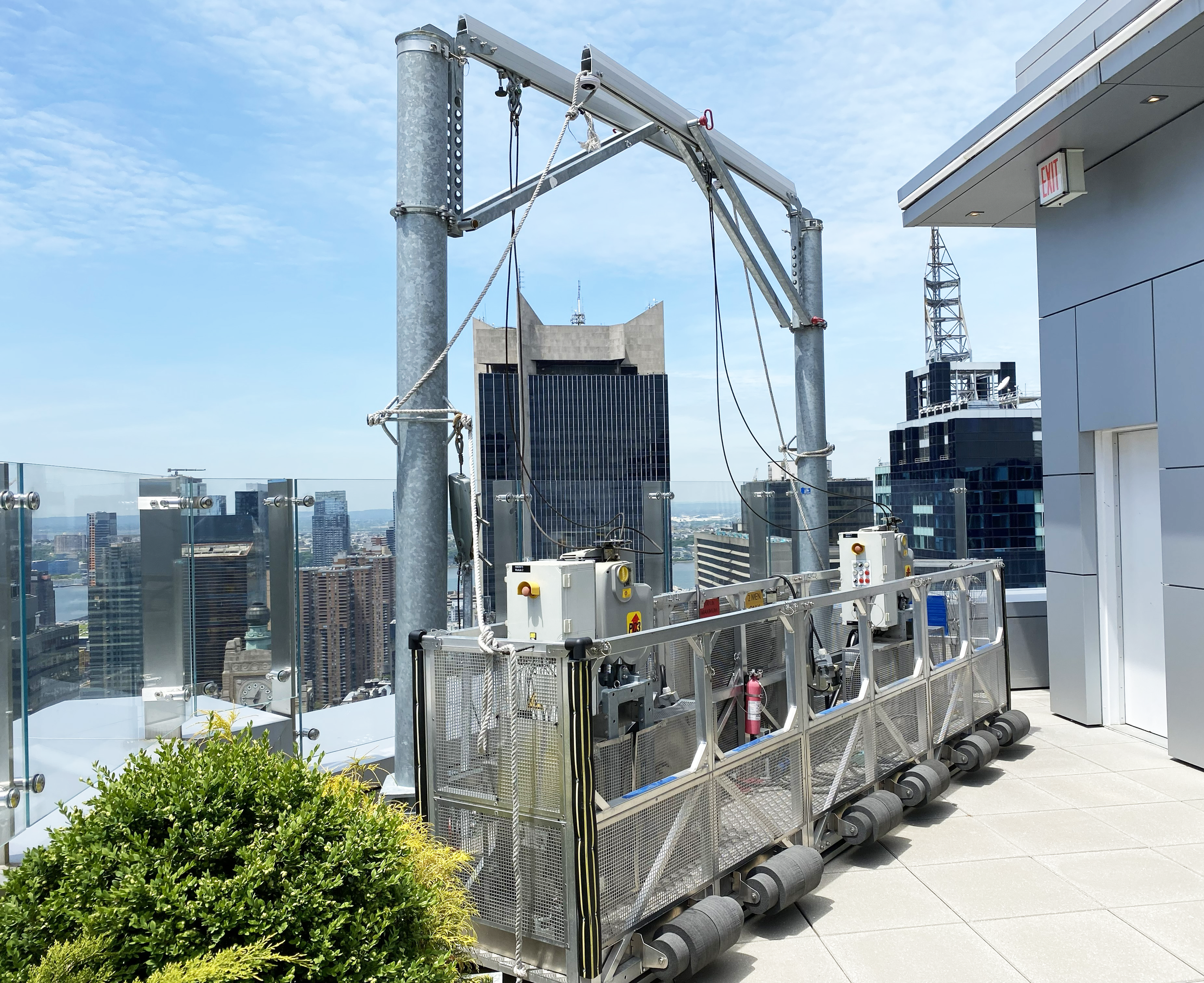 Rooftop cityscape view with a large metal elevator structure, glass barriers, and buildings in the background under a partly cloudy sky.
