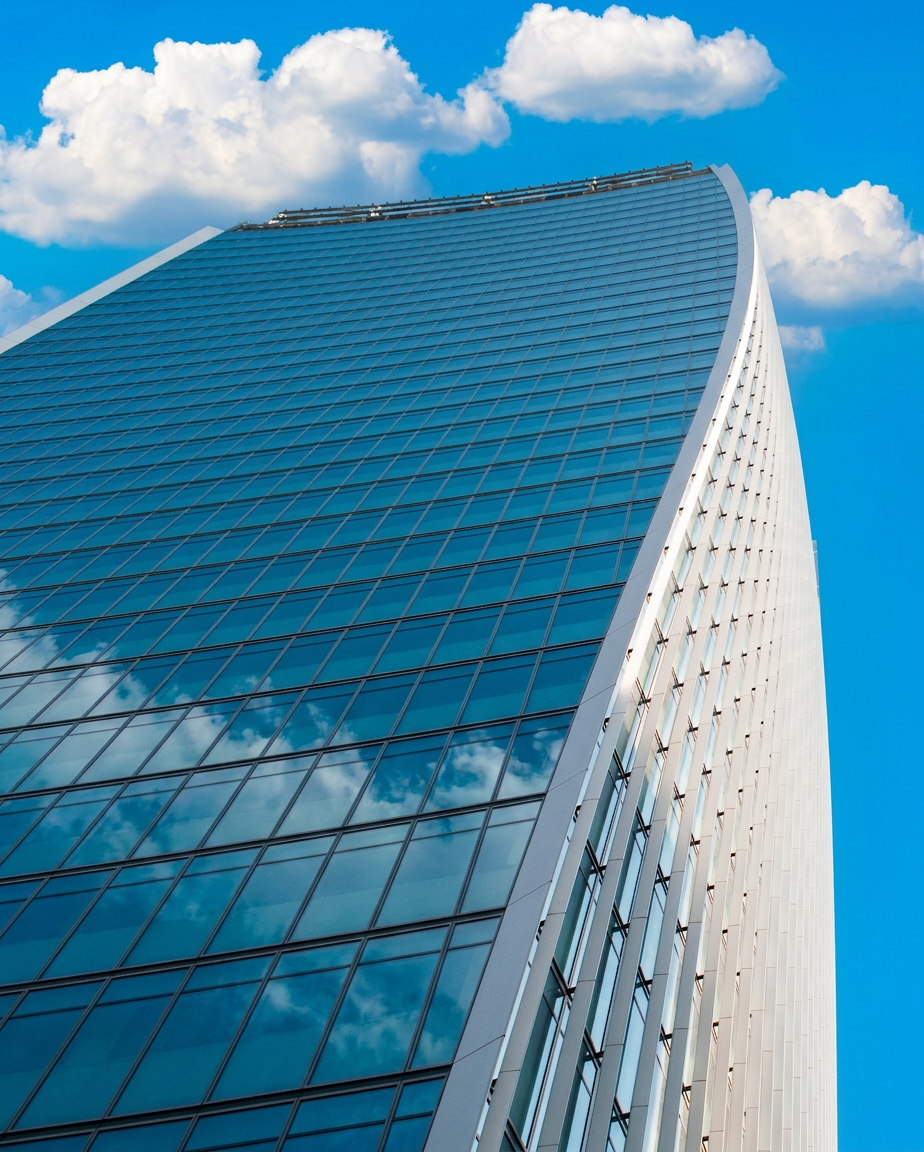 A tall modern glass skyscraper with a curved design, reflecting blue sky and white clouds.