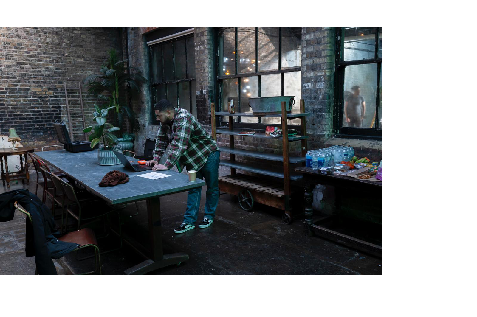 A man in a plaid shirt and jeans working on a laptop at a long table in an industrial-style room with exposed brick walls and large windows. There are bottles and snacks on a nearby table, and another person is visible outside through a foggy window.