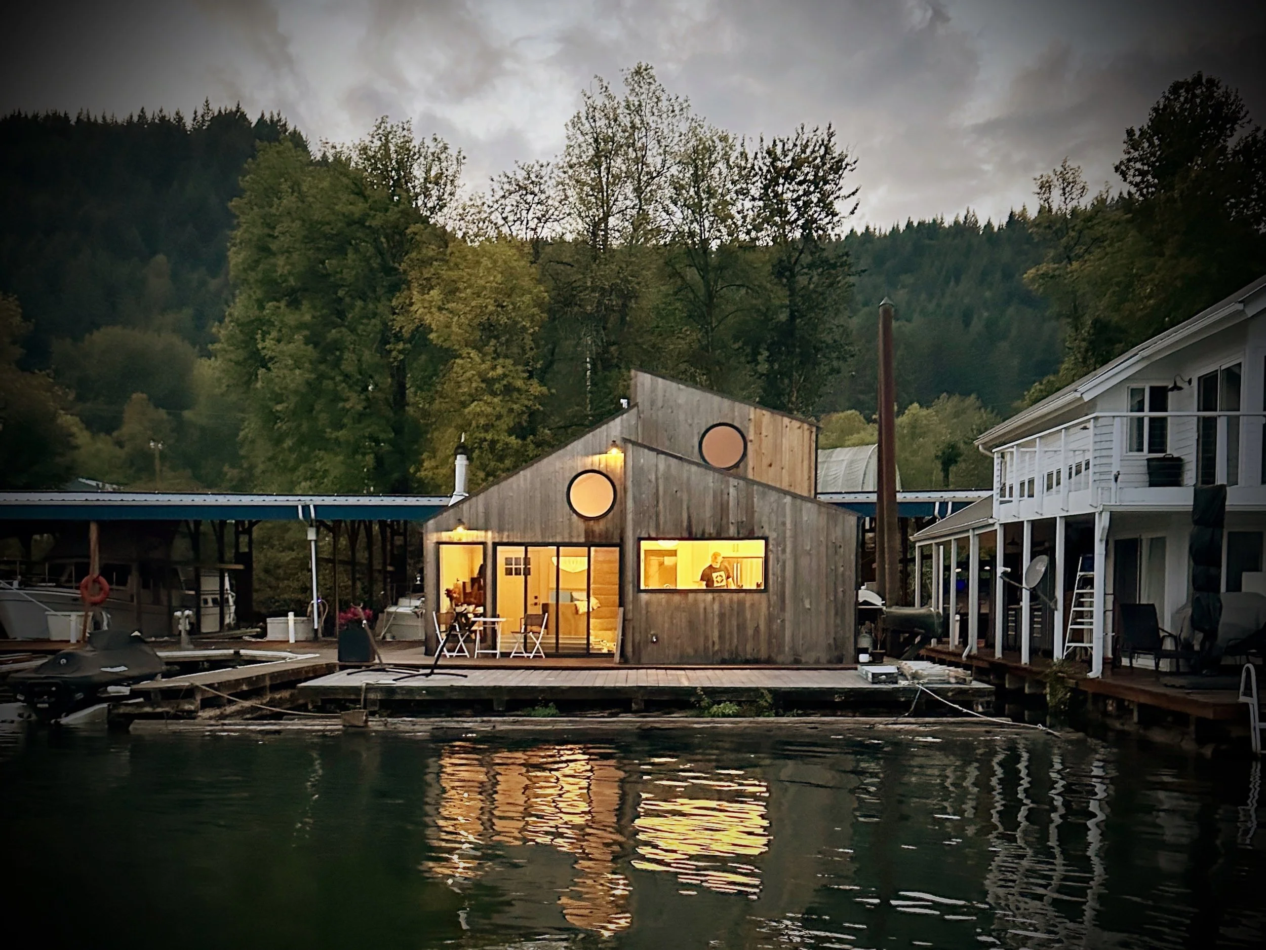 A lakeside scene at dusk with a house illuminated from within, surrounded by trees against a cloudy sky. The house has large windows and is near a deck extending over the water, with reflections visible on the lake's surface.