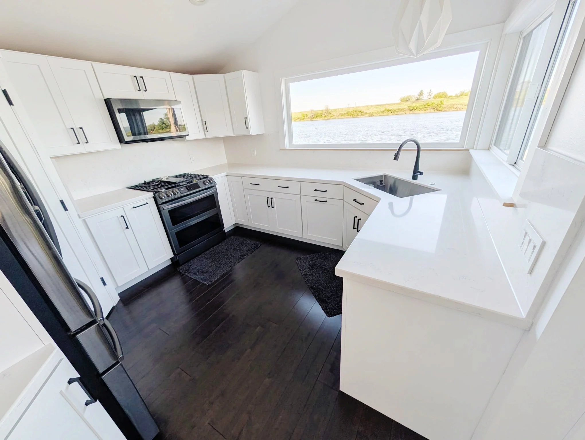 A modern kitchen with white cabinets, black handles, a black stove, and a large window overlooking a body of water and green trees in the distance.