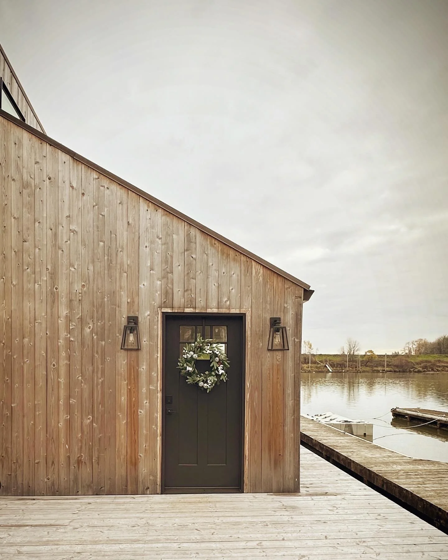 Wooden building by water with a door decorated with a holiday wreath, flanked by two lantern-style lights, with a dock and boats on the water in the background.