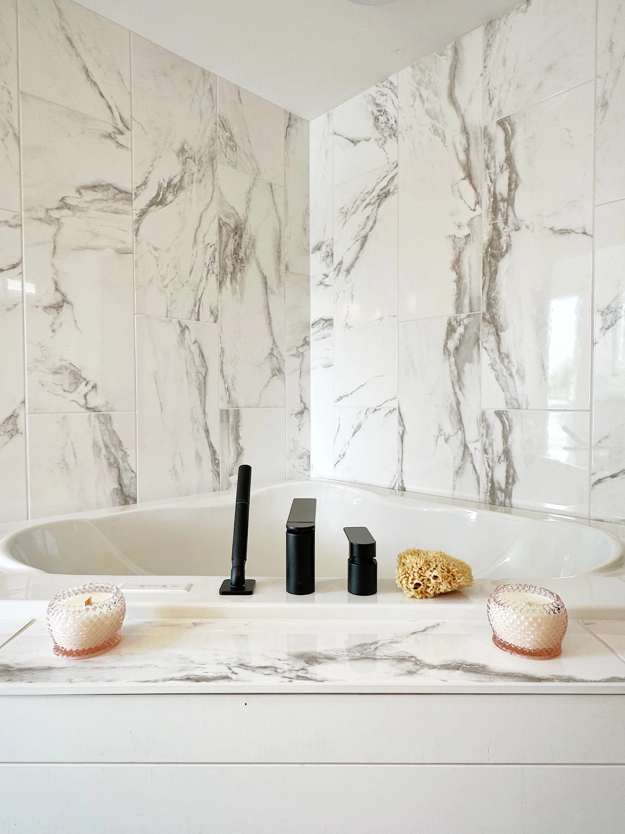 Bathroom with marble wall tiles, bathtub, black faucet, sponge, two pink candles, and decorative items.