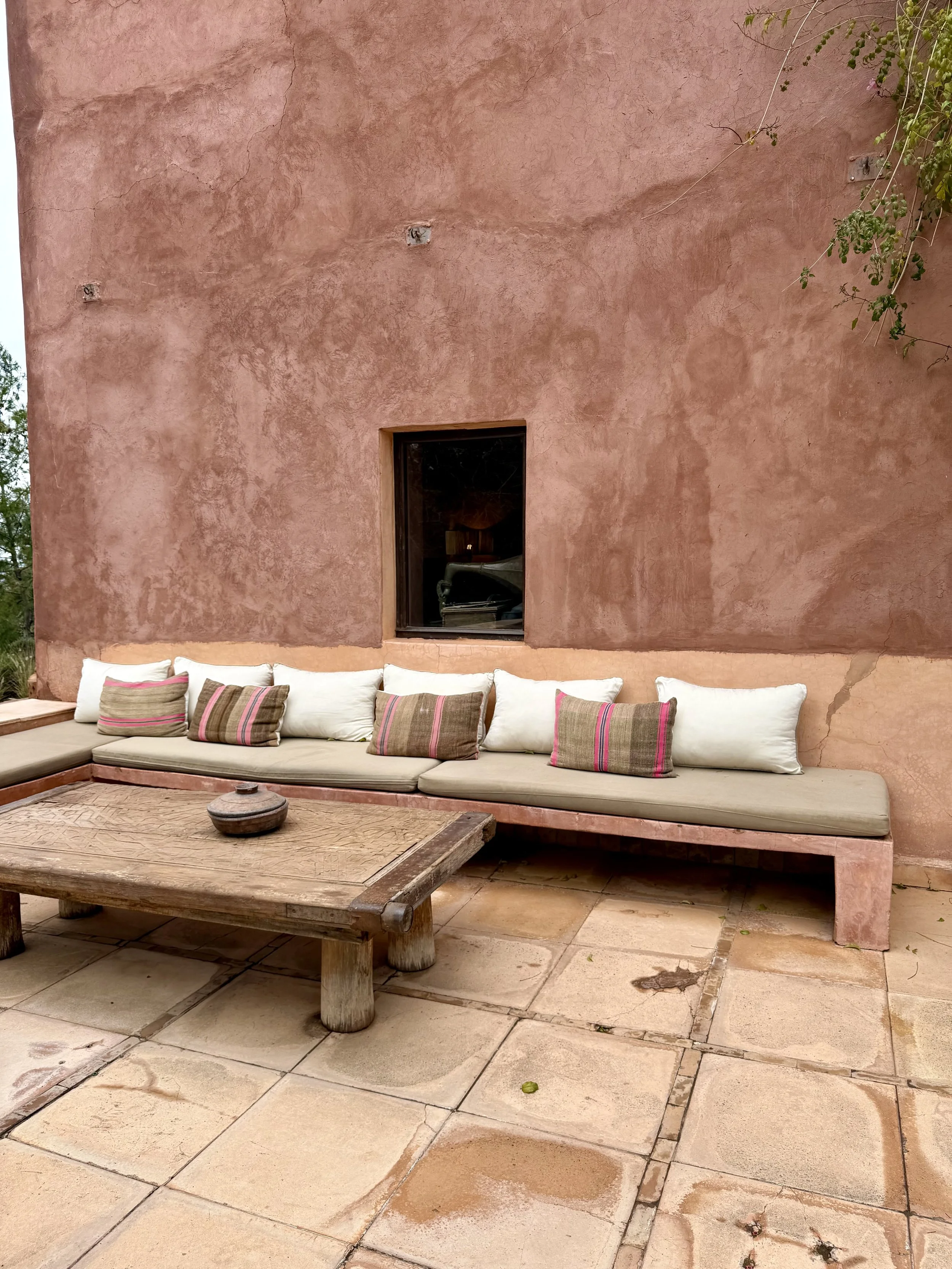A pink stucco wall with a small square window, surrounded by green ivy on the upper right. In front, there is a long beige bench with white and brown striped pillows, and a rustic wooden table on a tiled patio floor. Atlas mountains morocco