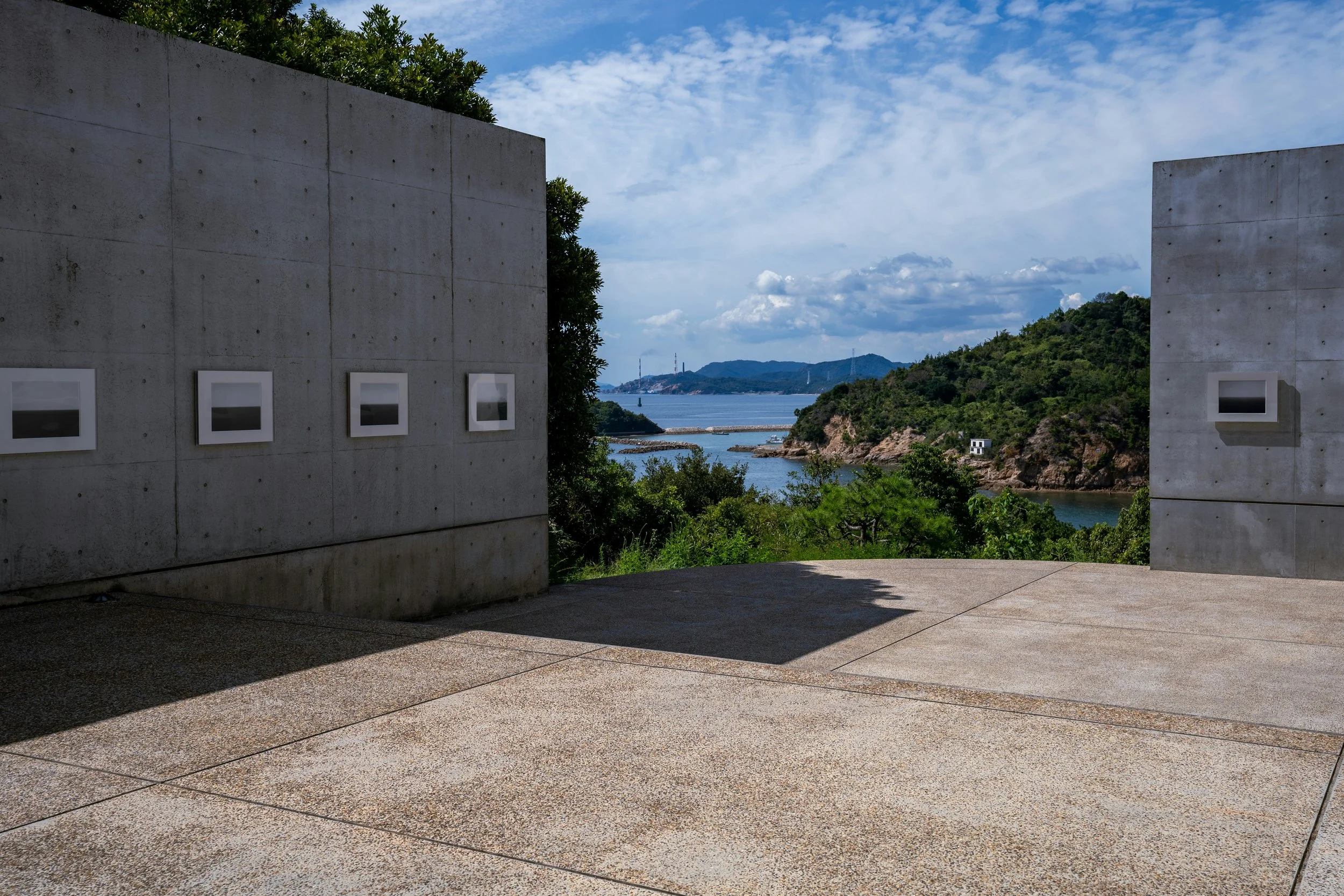 Framed black-and-white seascape photographs mounted on Tadao Ando's concrete walls at Benesse House, with a view to the green hills and the Seto Inland Sea beyond.
