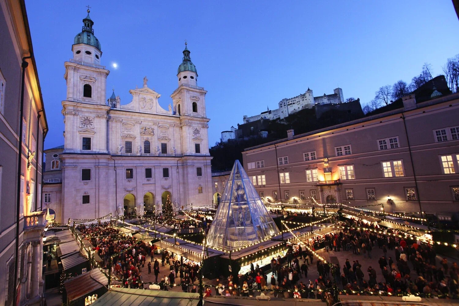 Salzburg's Christkindlmarkt on Domplatz at dusk, with the cathedral and Hohensalzburg fortress lit above the market stalls.